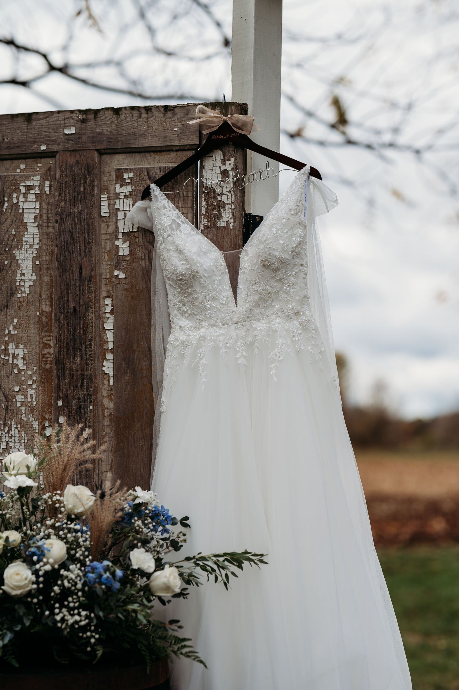 A white wedding dress hangs against a rustic wooden wall with delicate floral arrangements featuring blue and white flowers.