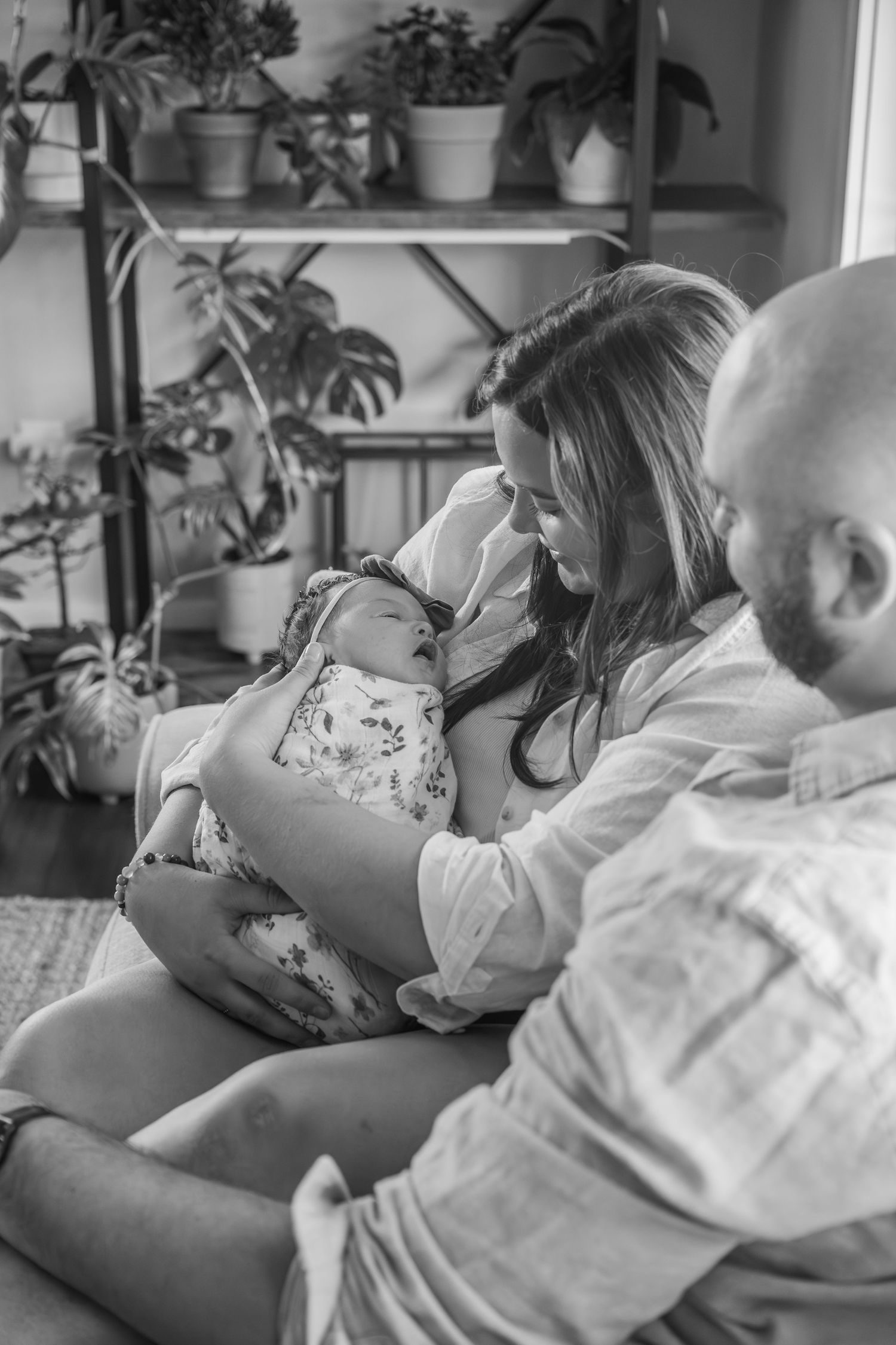A tender black and white moment in a room with potted plants on shelves, showing a family with a small baby.