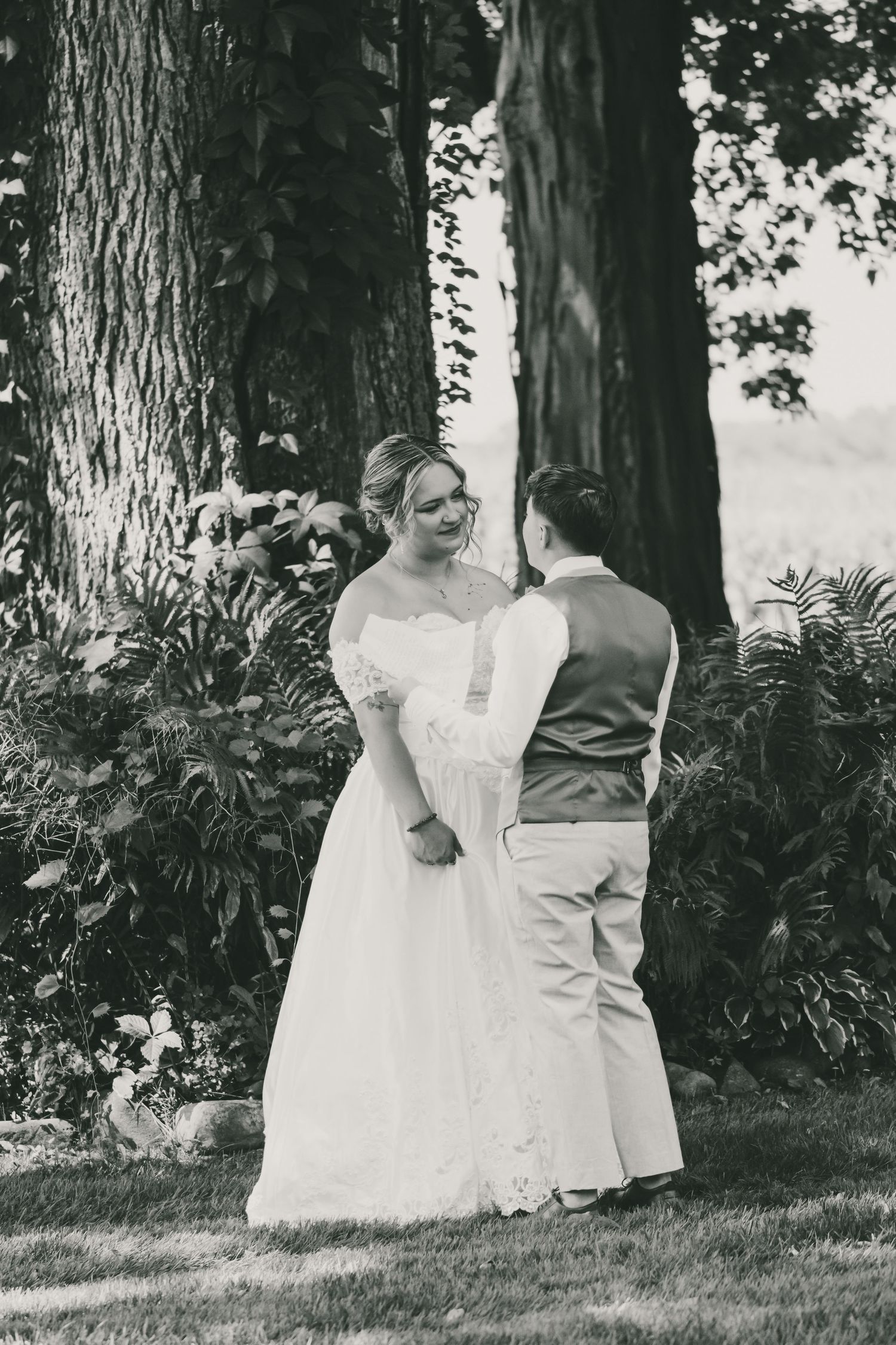 A black and white photo series shows a couple in wedding attire walking together among trees and ferns in a garden setting.