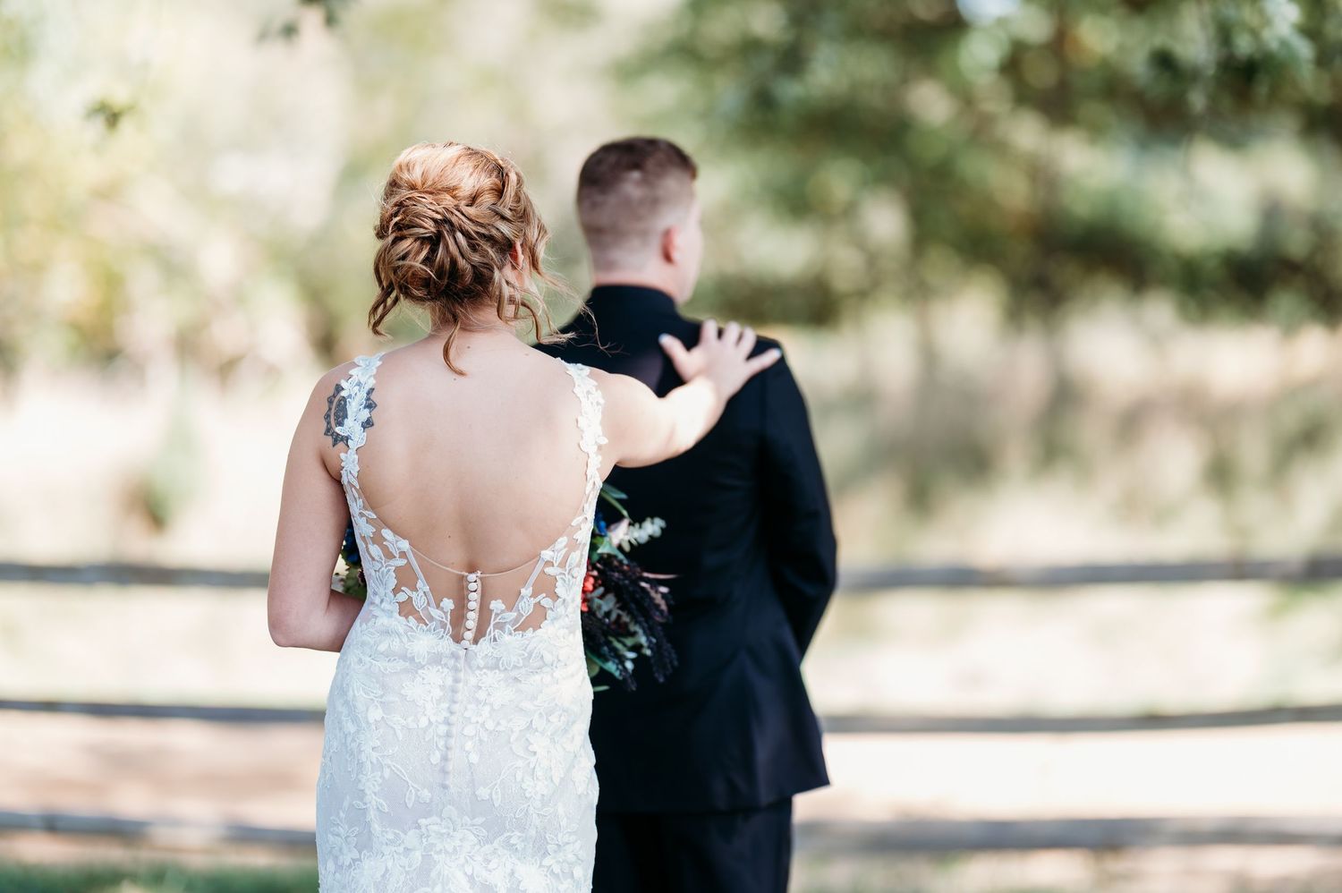 A couple in wedding attire stands outdoors, with the bride wearing a detailed backless lace gown.