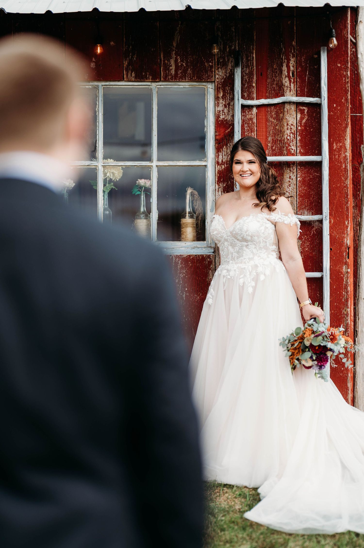 A bride in a flowing white gown poses in front of a rustic red barn during a wedding photoshoot.