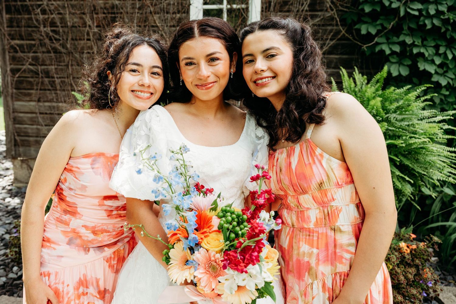 Bride in white gown holds colorful bouquet flanked by two bridesmaids in coral floral dresses outdoors.