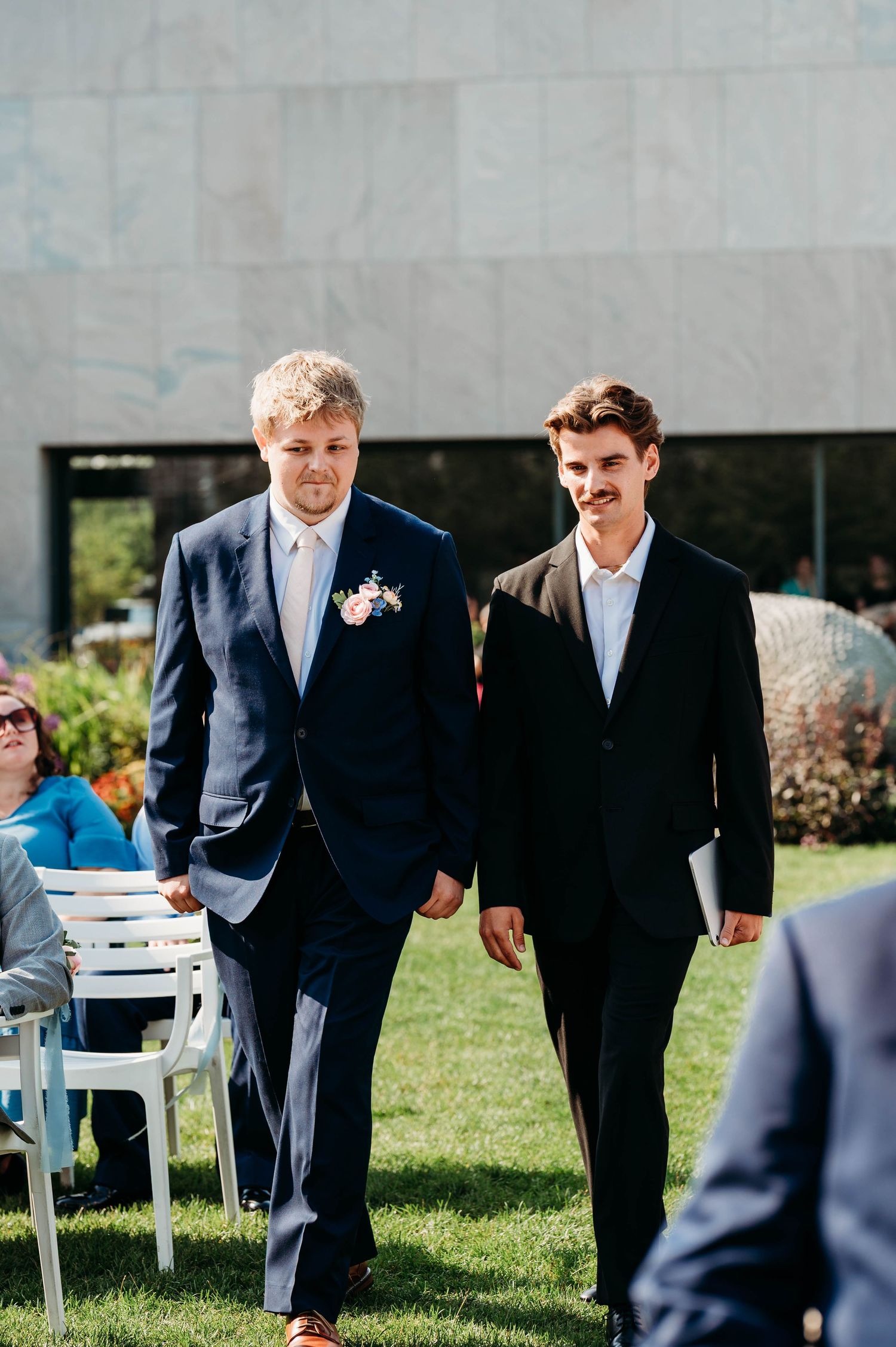 Two grooms walk hand in hand down the aisle at an outdoor wedding ceremony on a sunny day.