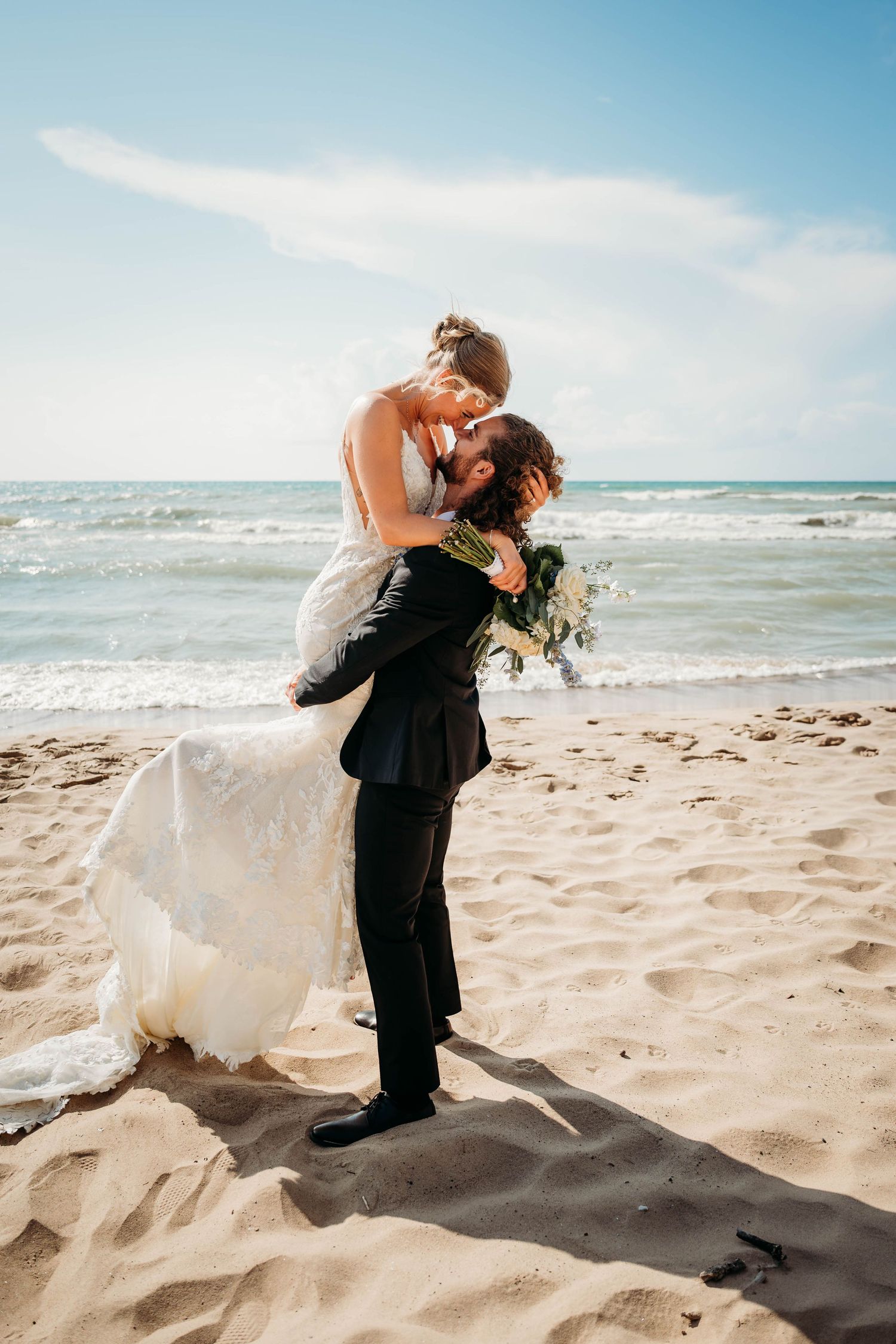 Groom lifts bride in white wedding dress for a romantic beach kiss, with ocean waves and blue sky backdrop.