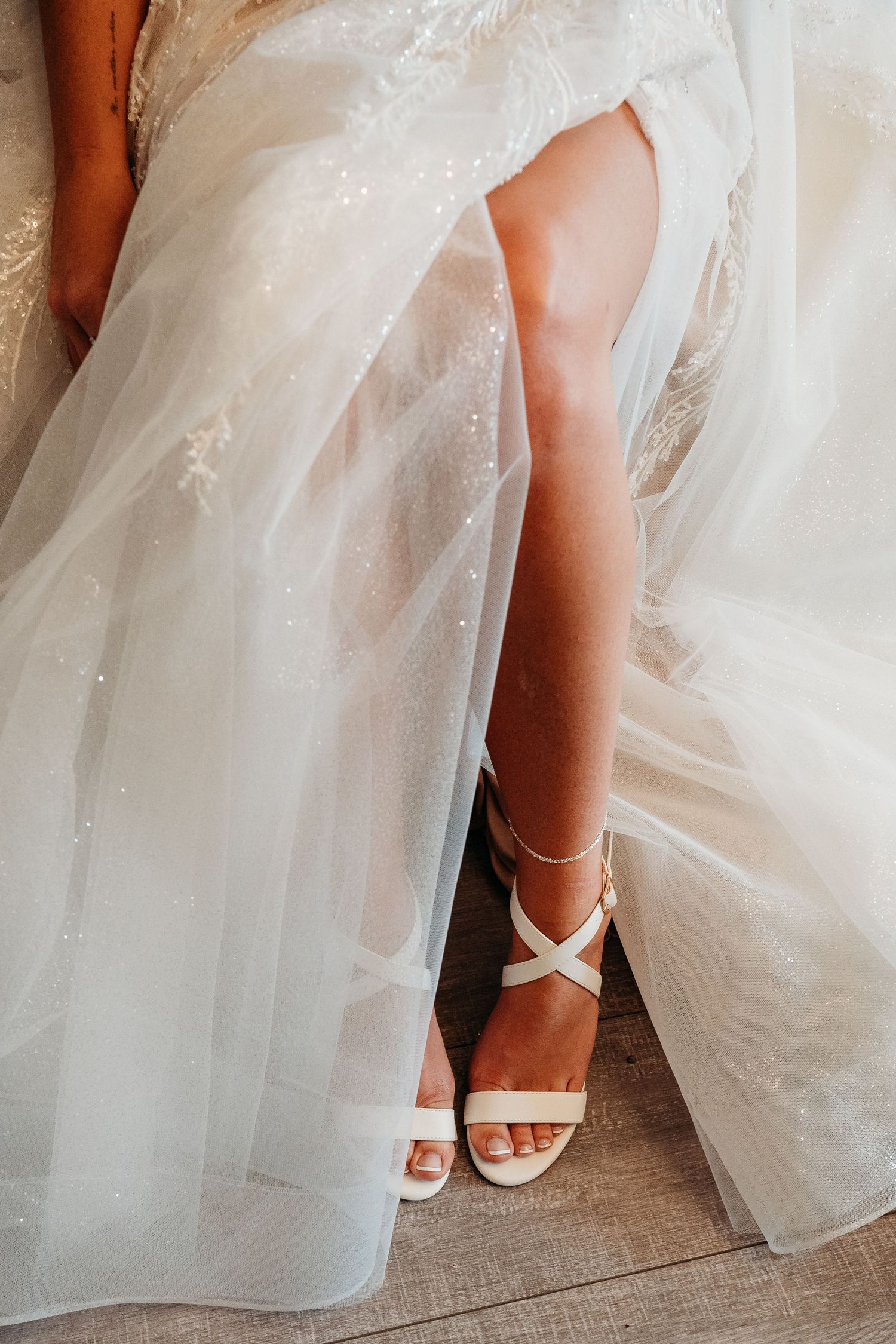 White strappy bridal sandals peek out from beneath layers of tulle and lace wedding dress fabric.