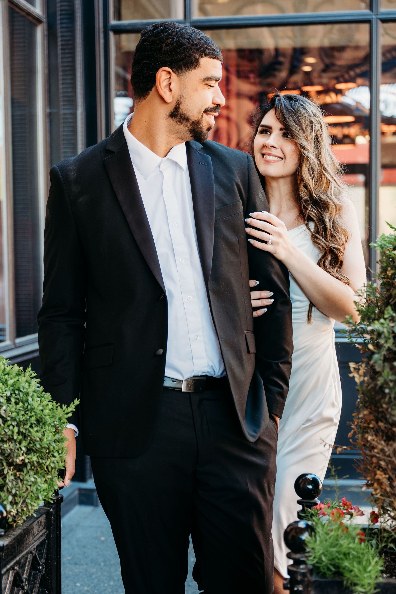Couple in formal attire sharing a romantic moment outside a building with green potted plants.