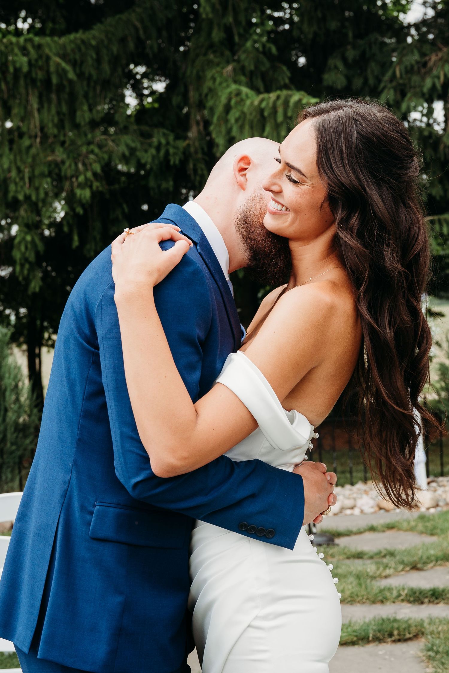 A romantic wedding couple embraces outdoors in a tender moment against a natural backdrop of green trees.