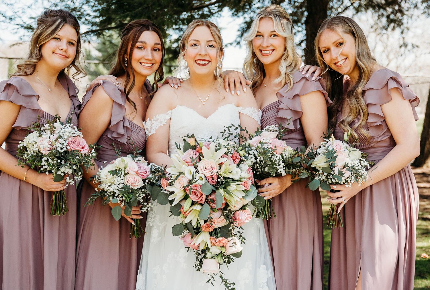 Group of bridesmaids in dusty rose dresses hold pink and white bouquets during outdoor wedding celebration.