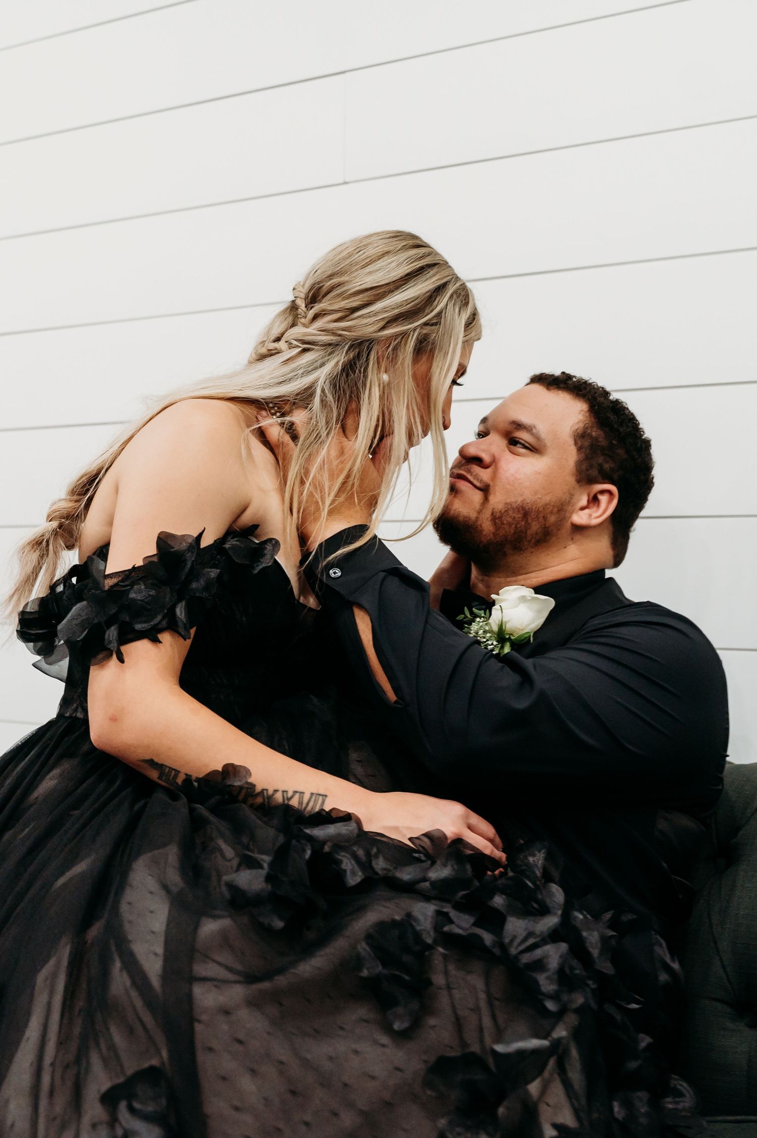 A couple shares a romantic moment outdoors in coordinated black outfits against a white wall.