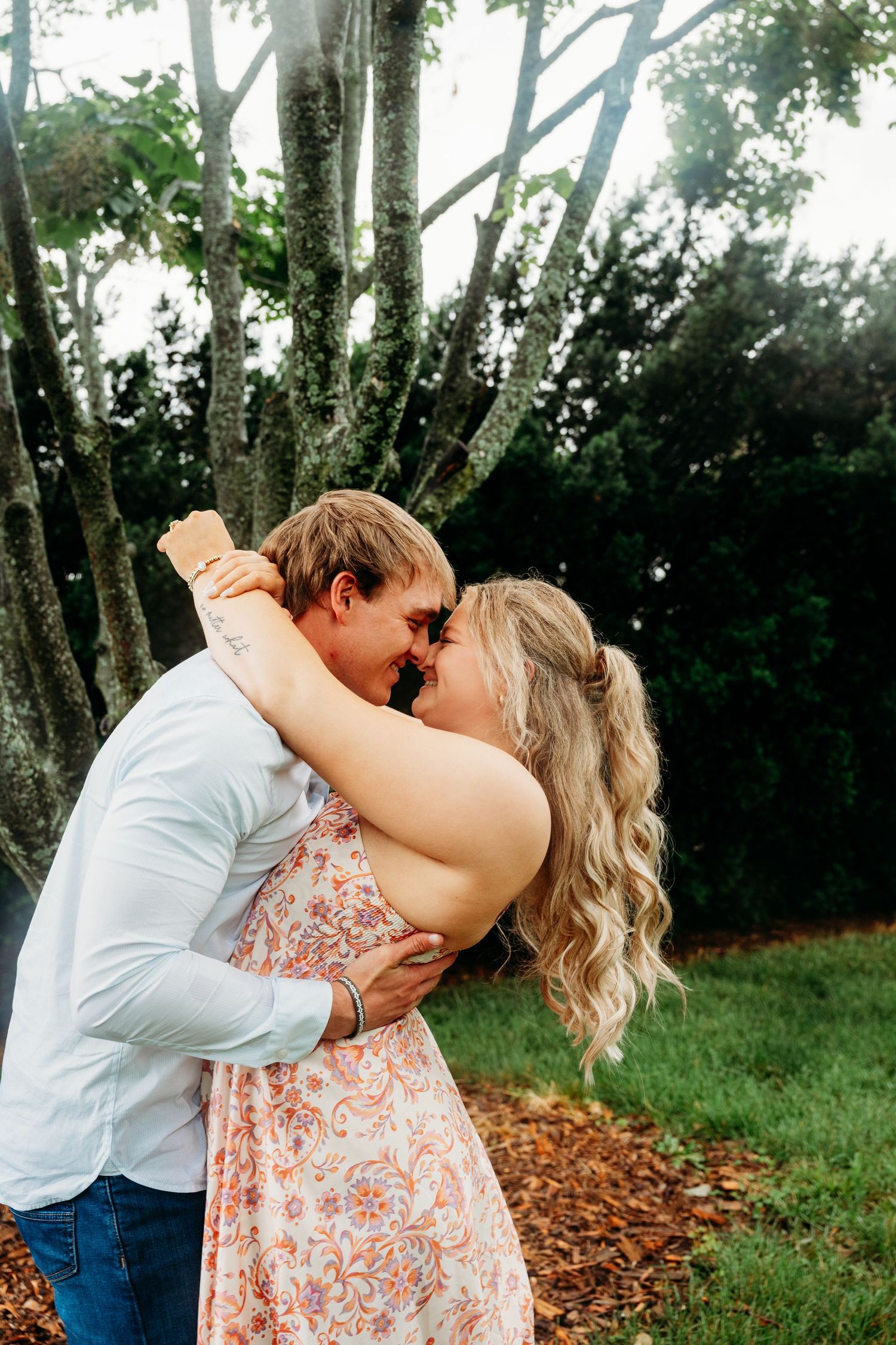 Couple sharing a romantic forehead touch embrace outdoors surrounded by lush green trees and fallen leaves.
