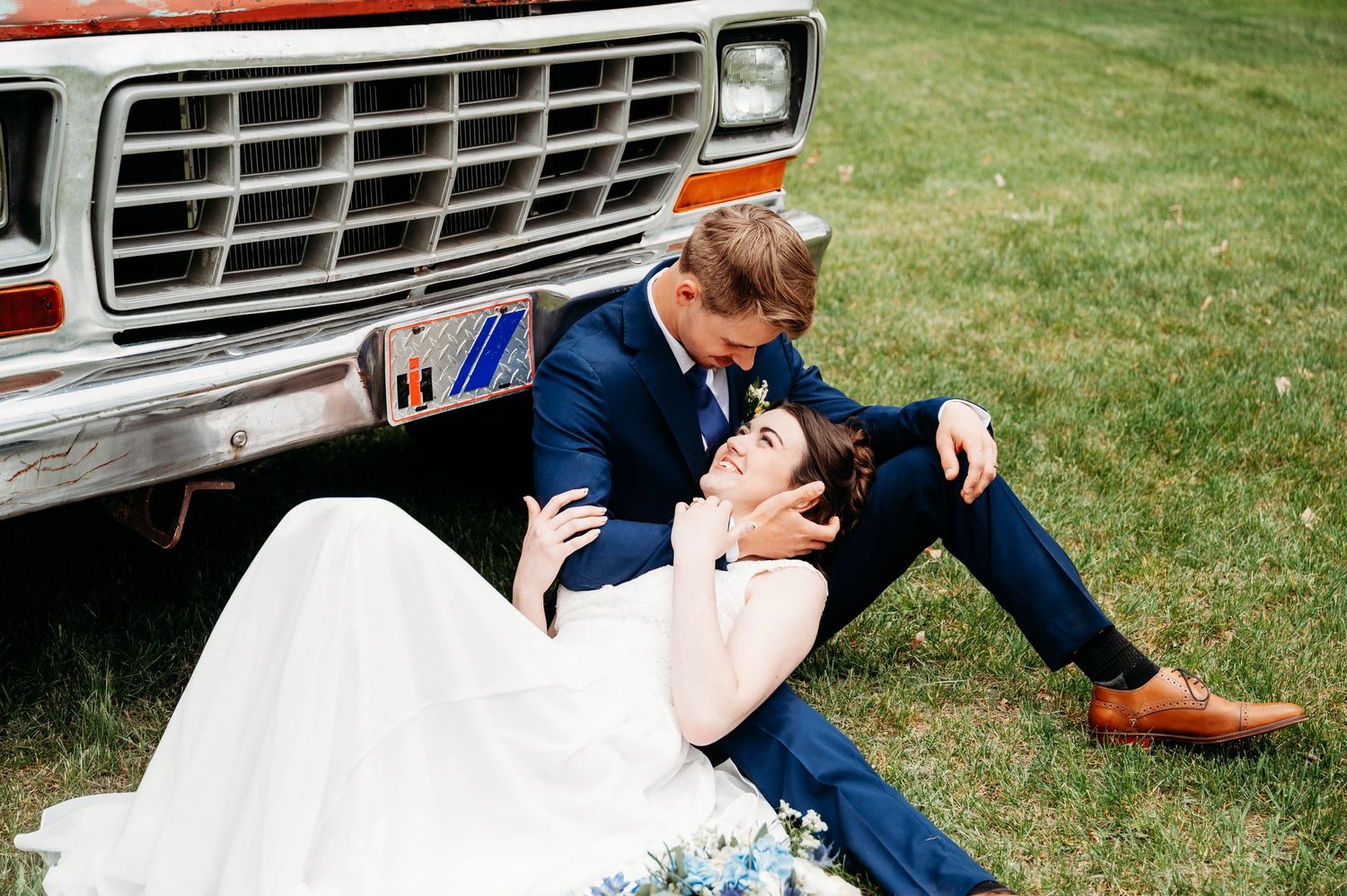 Bride and groom sitting together on grass leaning against vintage white truck during outdoor wedding photo session.