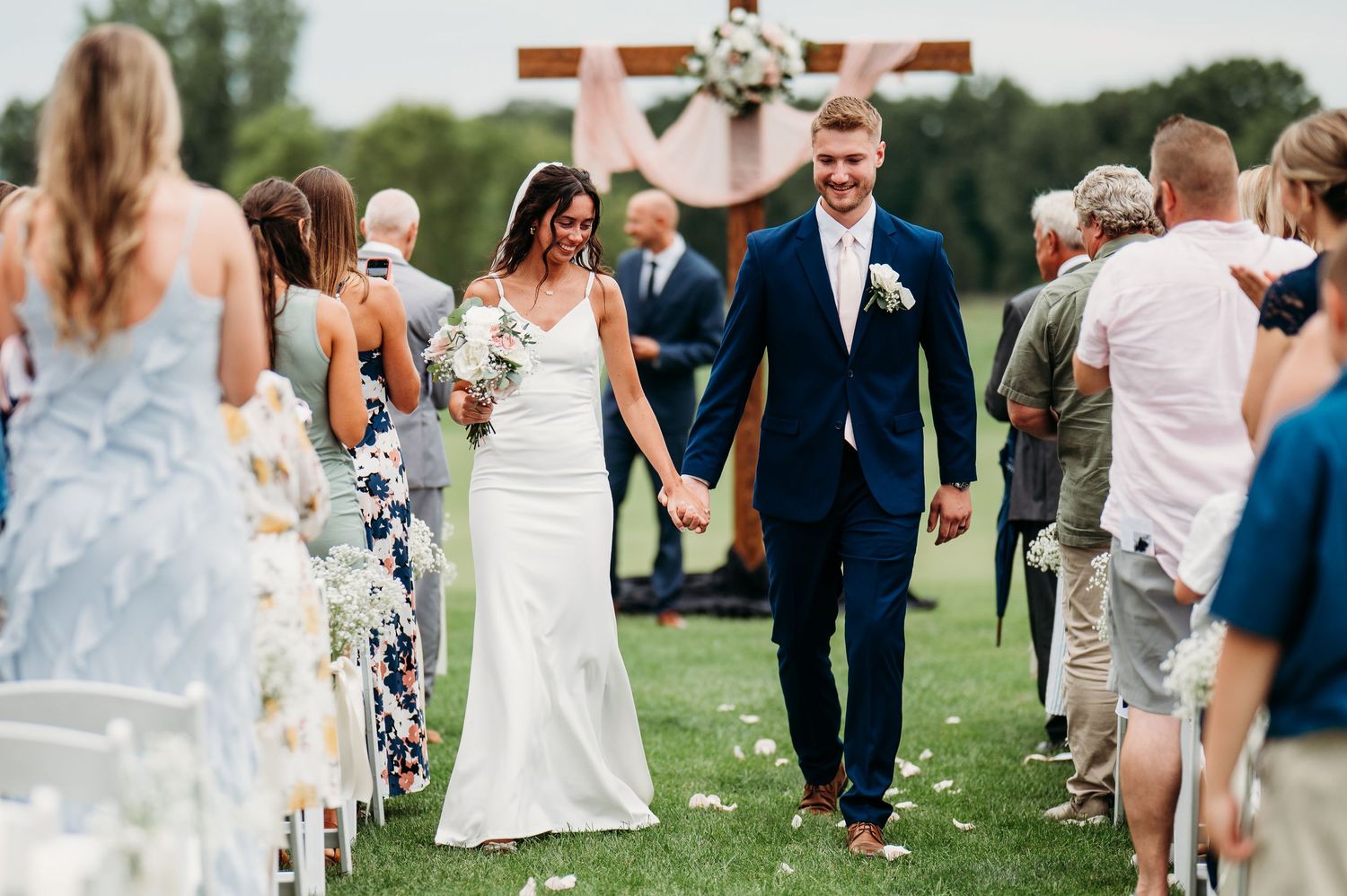 Bride and groom walk hand in hand down outdoor aisle after wedding ceremony with floral cross backdrop.