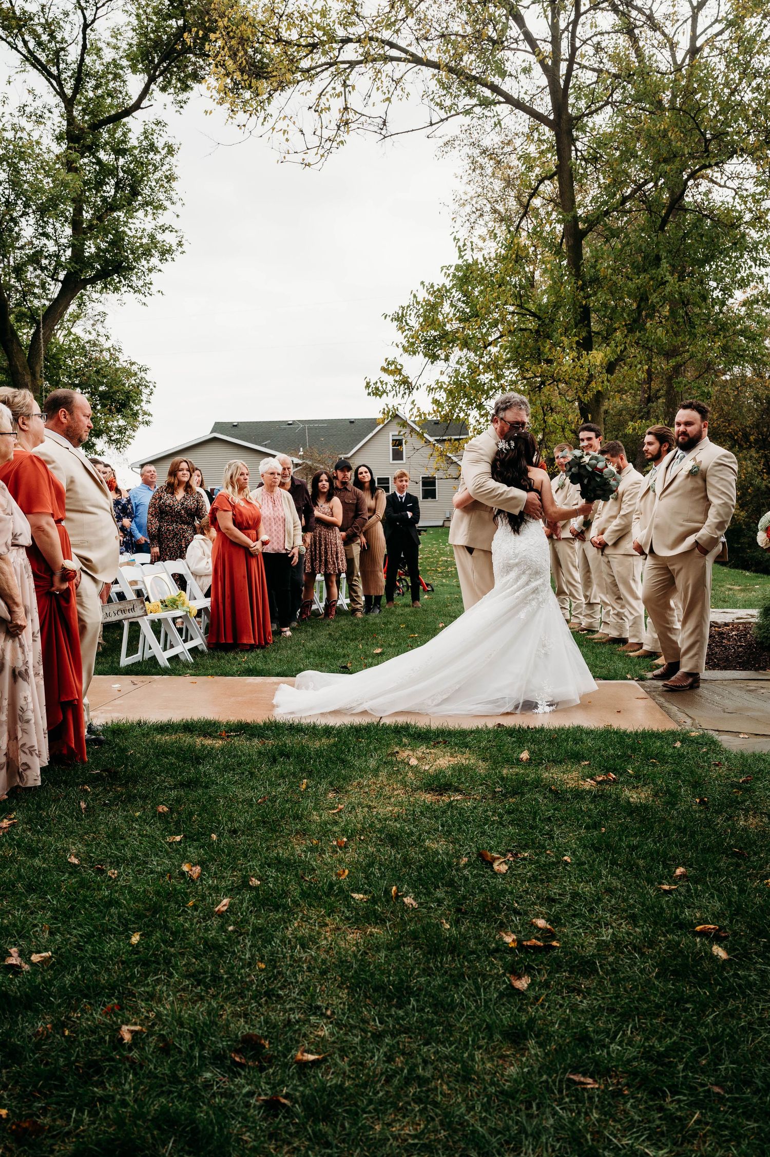 Outdoor wedding ceremony with bride and groom sharing first kiss on grass aisle, guests watching, bridal party in tan suits and red dresses.