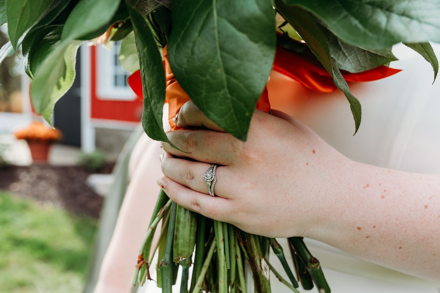 Close-up of a bride holding a green bouquet with an orange ribbon, showcasing a diamond wedding ring on her finger.