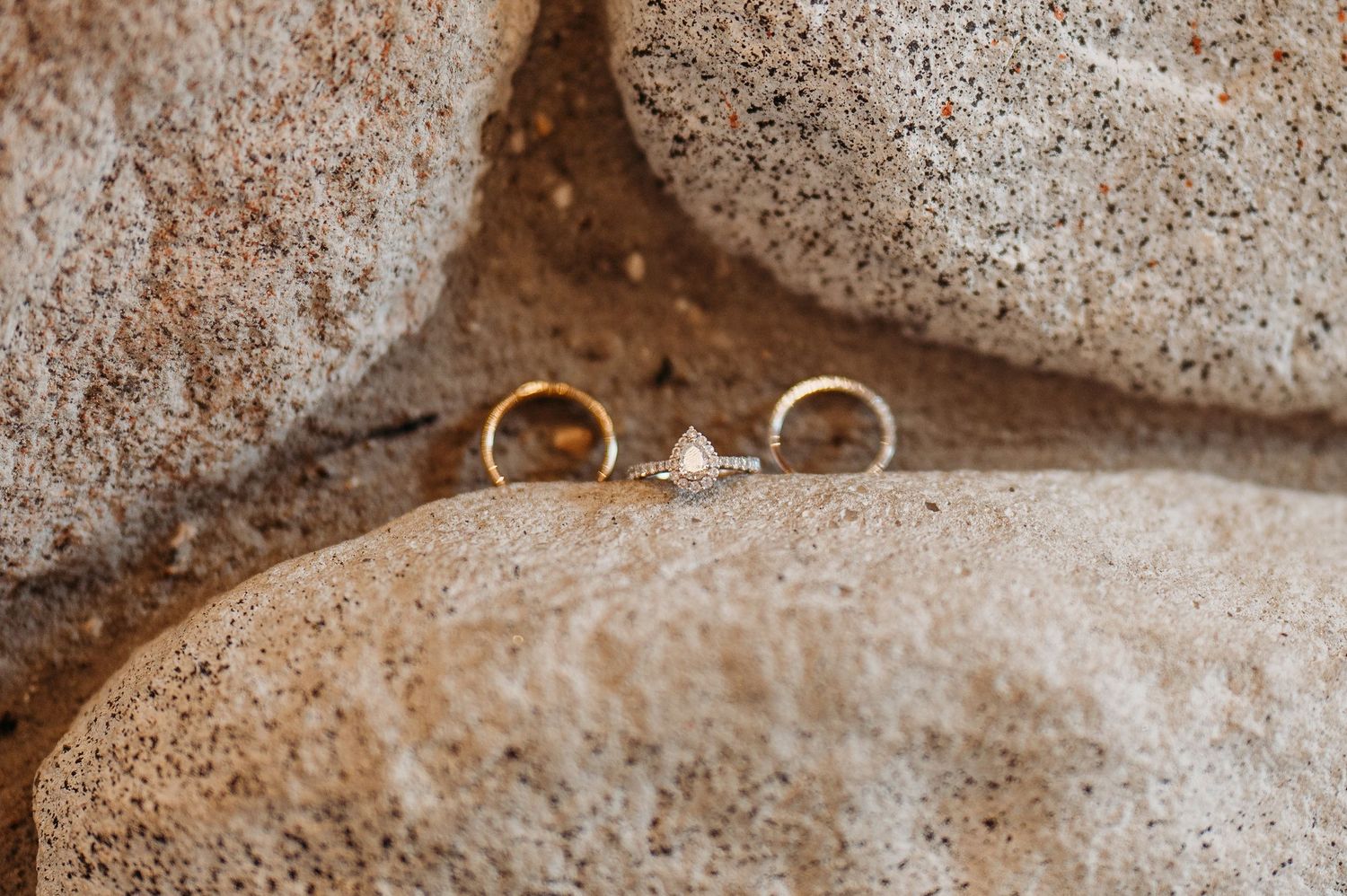 Two gold wedding bands and a diamond engagement ring displayed on rustic stone rocks outdoors.