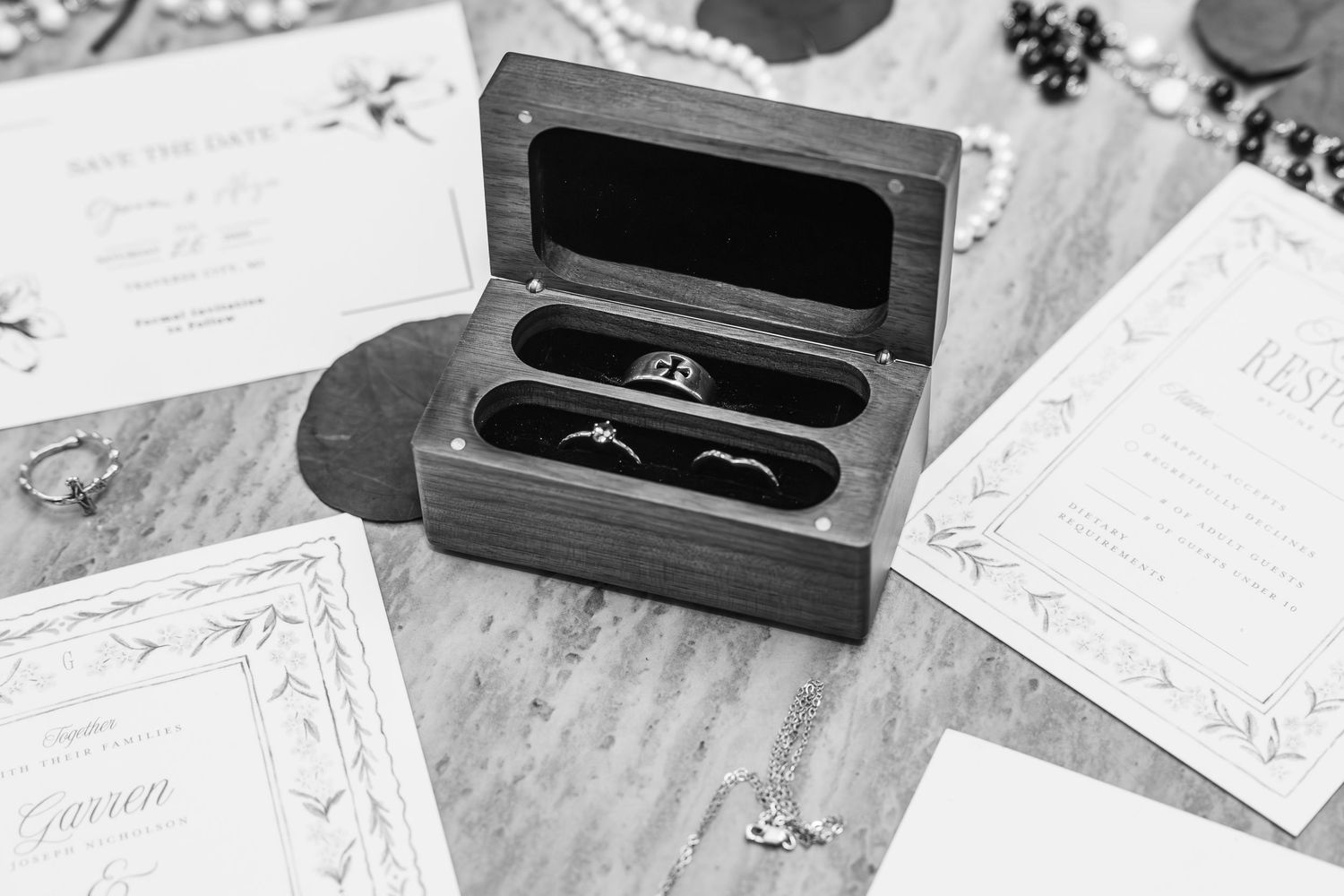 Black and white wedding flat lay with wooden ring box holding two bands, engagement ring, invitations, and jewelry on wood surface.