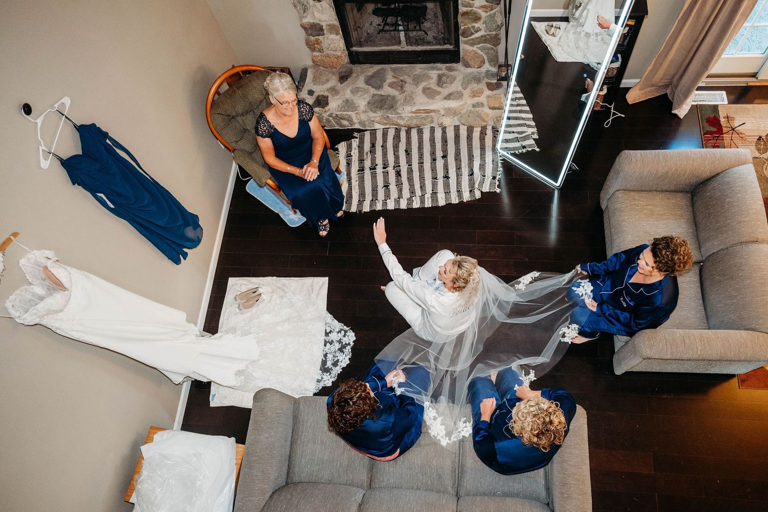 Aerial view of bride in white wedding dress surrounded by bridesmaids in navy blue robes during bridal preparation.