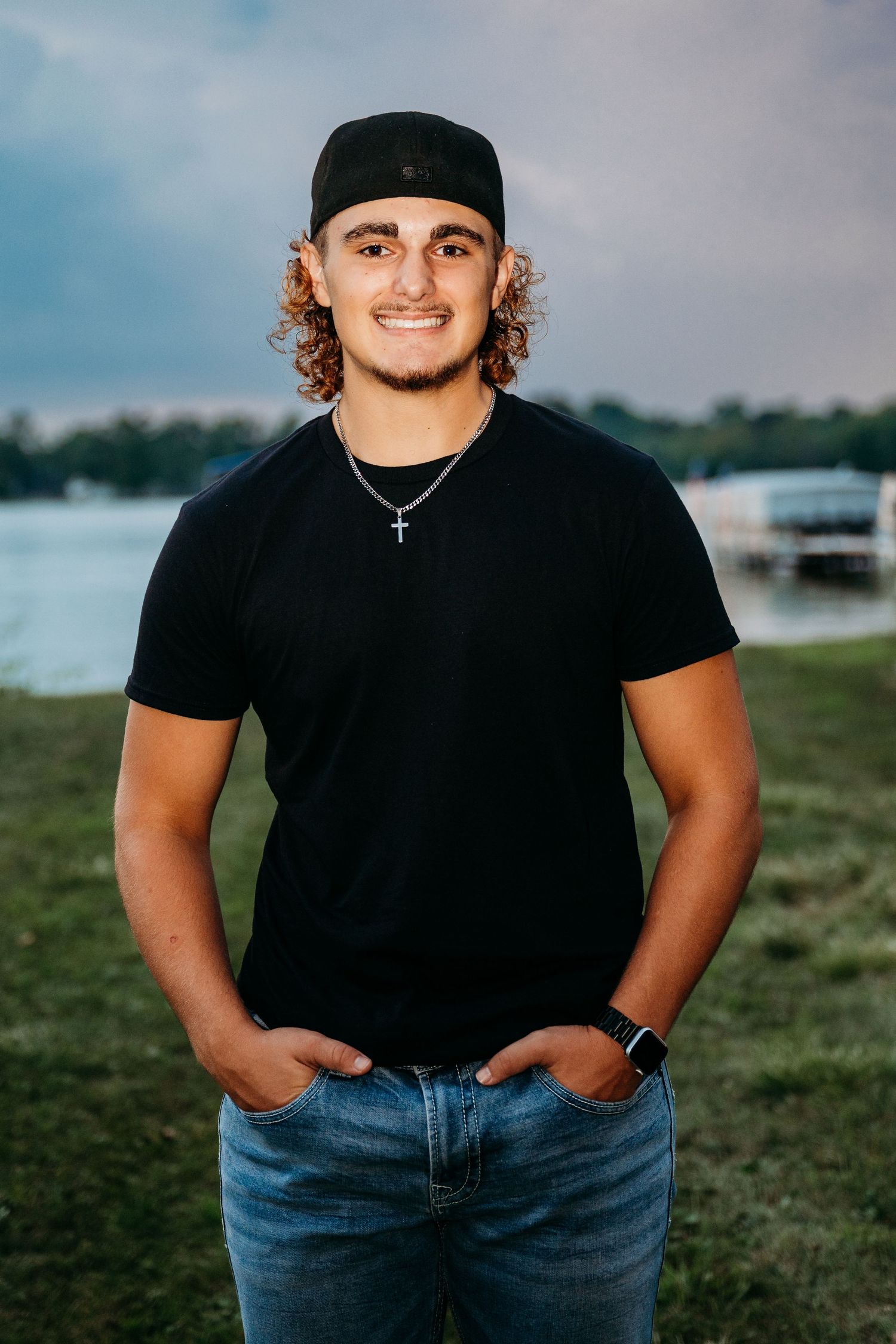 Person wearing black t-shirt and jeans poses casually outdoors in front of water on grassy area.