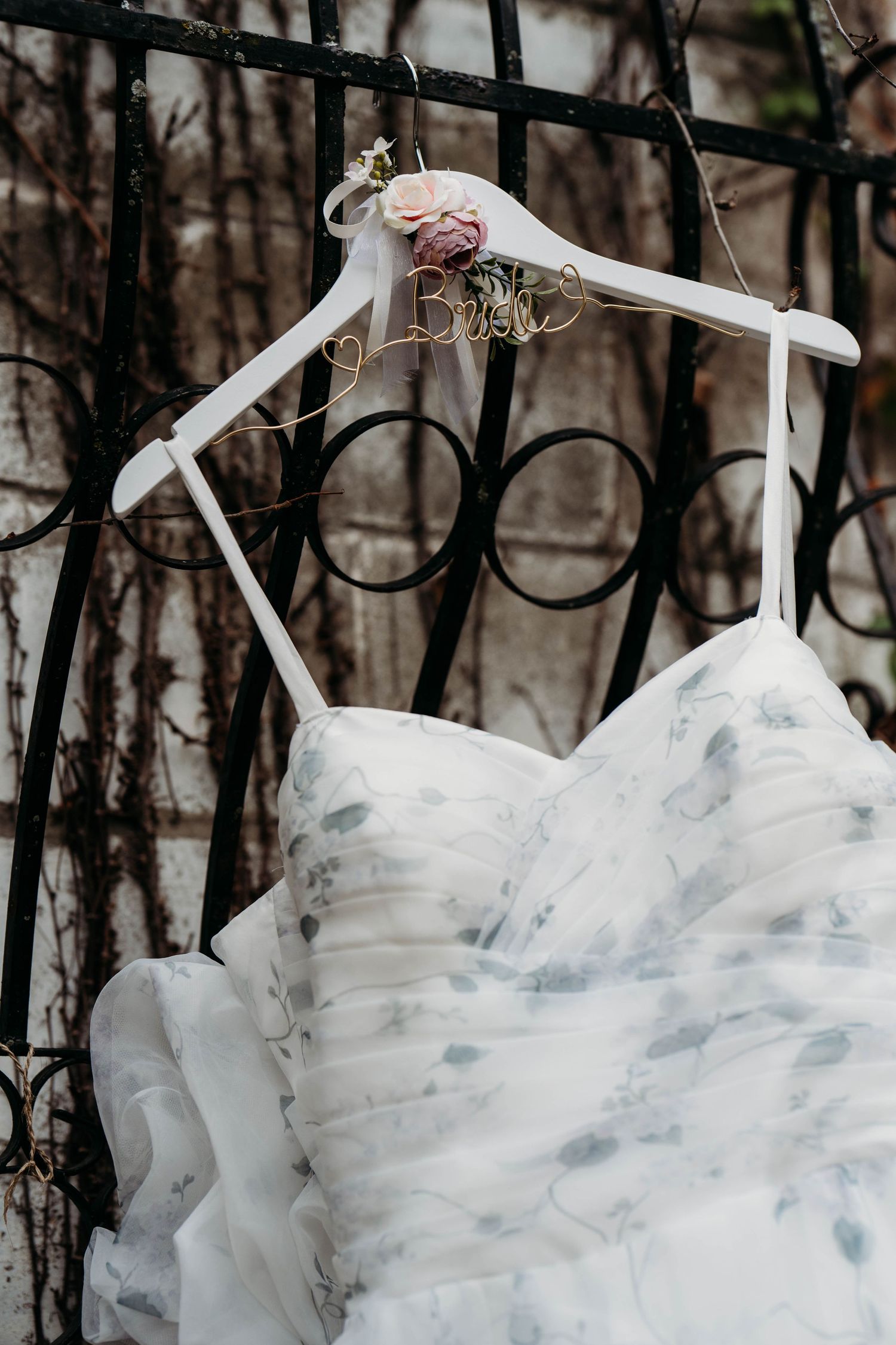 A delicate white wedding dress hangs on a decorative hanger against an iron gate with vines in the background.
