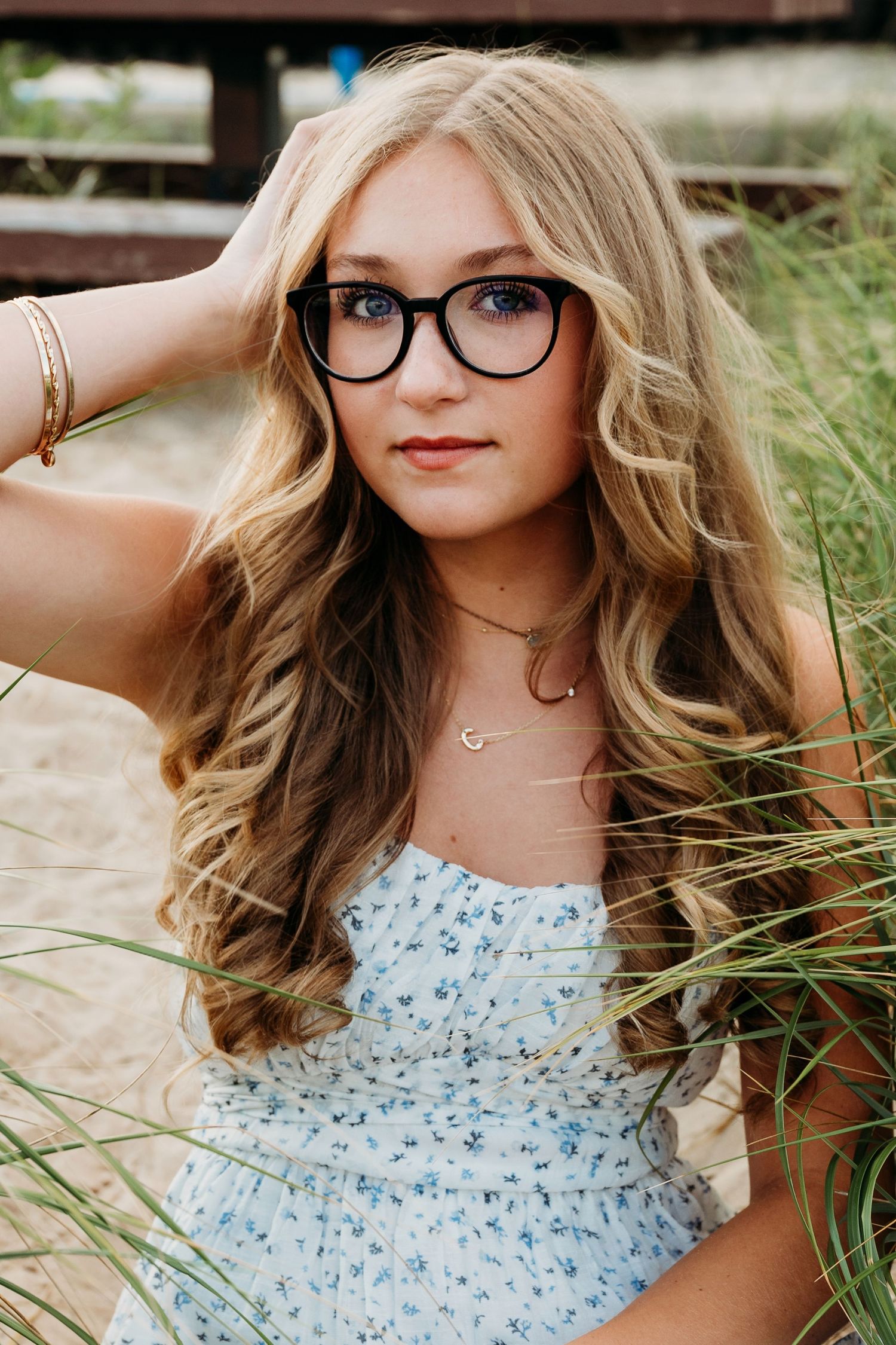 Young woman in white floral dress and glasses poses in tall grass against cloudy sky.