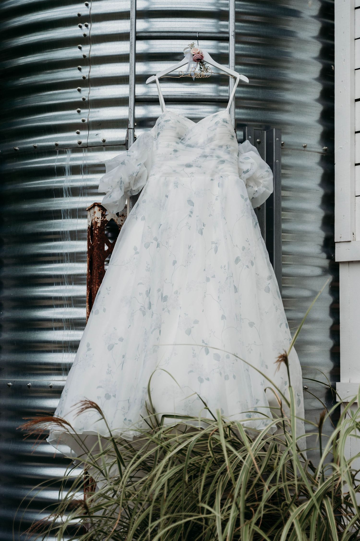 A white wedding dress hangs on a hanger against a corrugated metal silo wall with ornate floral details and grass below.