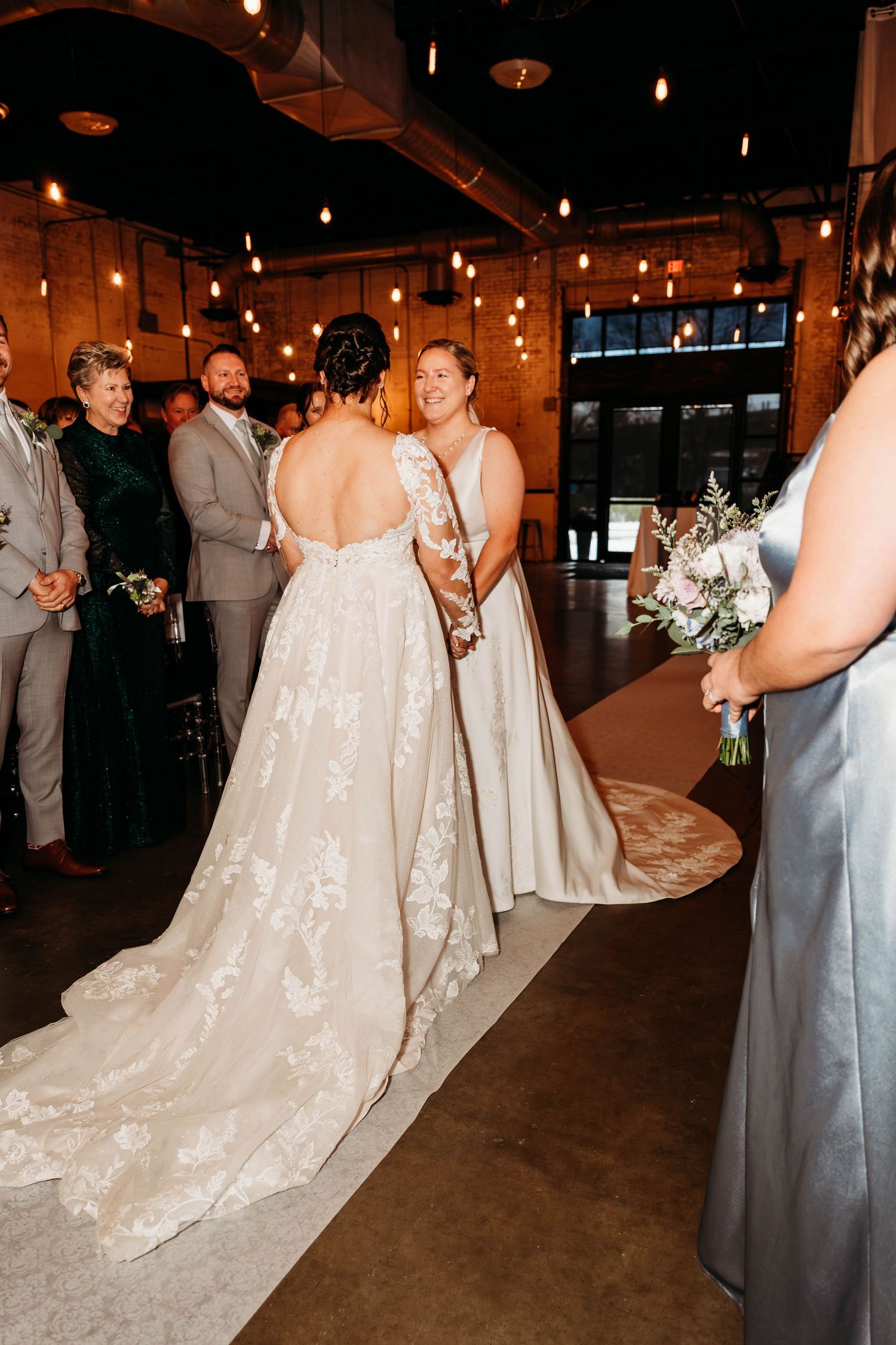 Wedding party in formal attire gathers under warm lighting in an industrial reception venue.