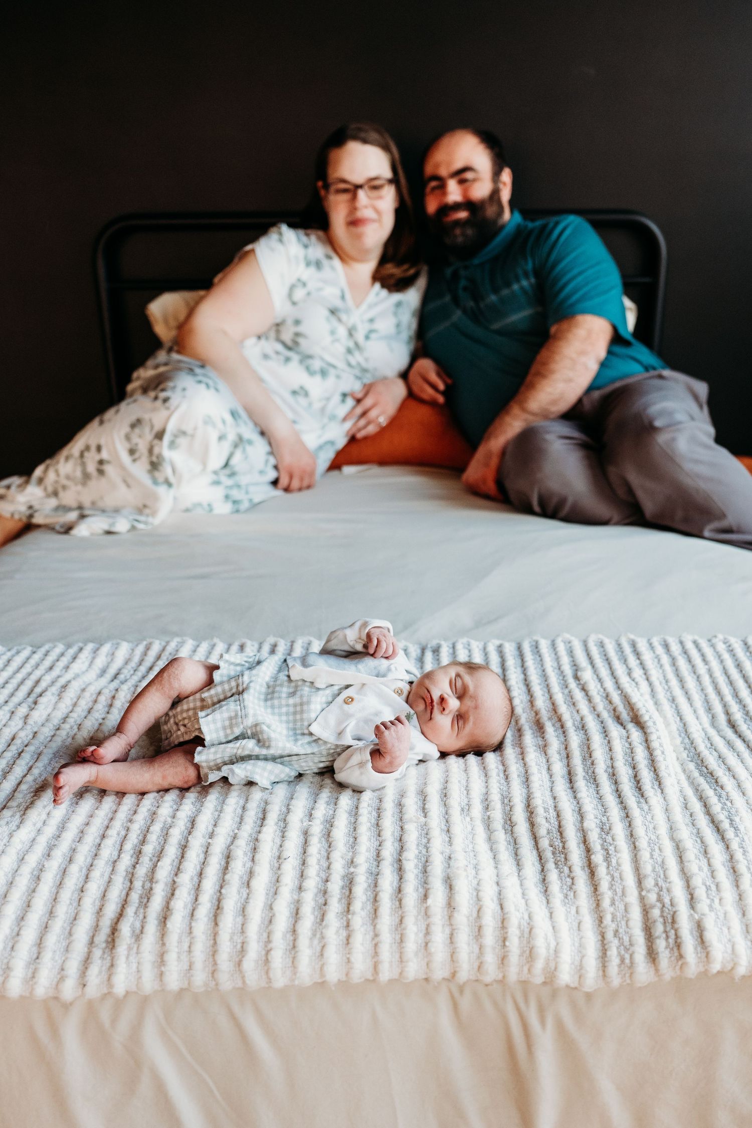A couple relaxes on a bed with their baby in a floral dress, creating an intimate family portrait series.