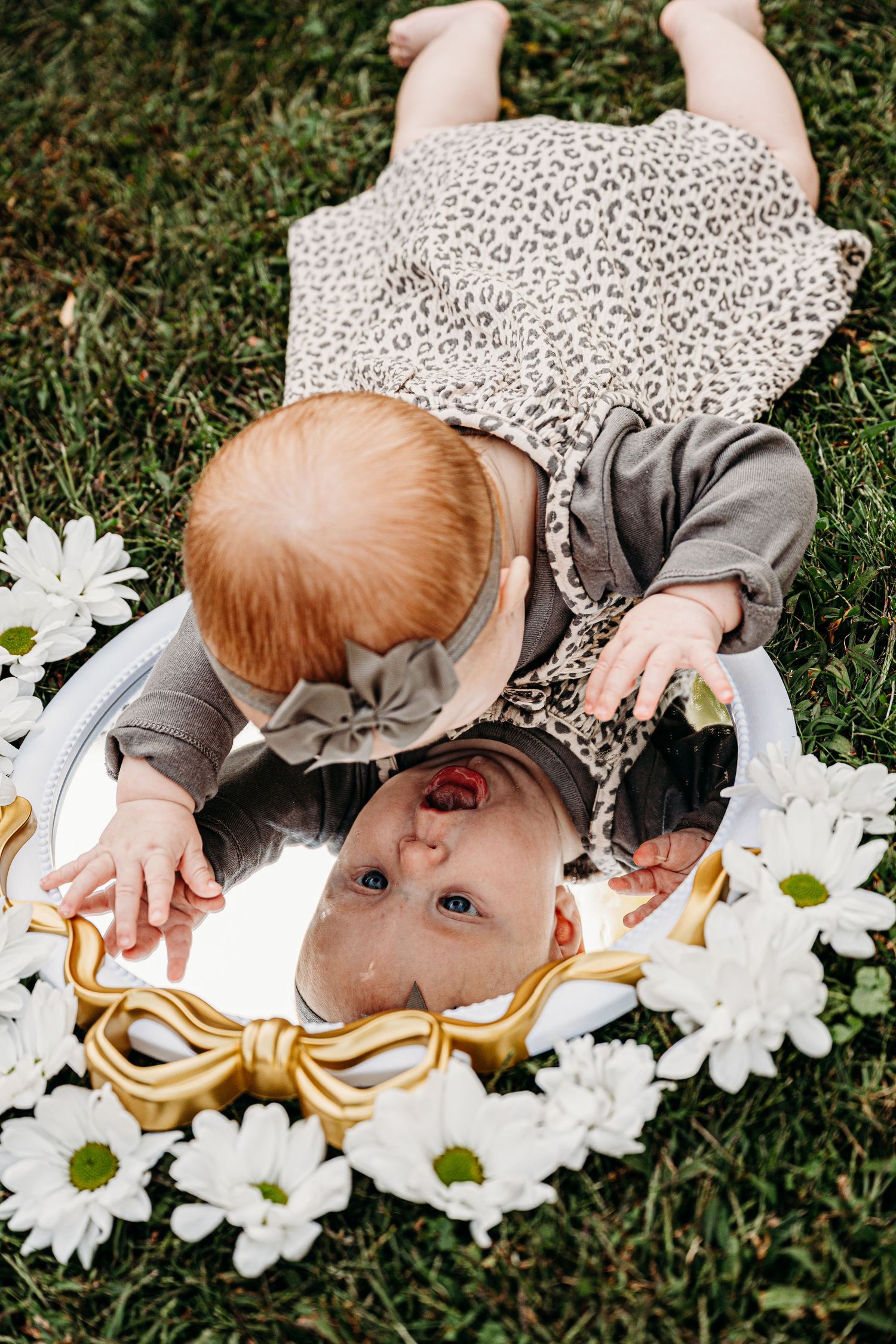 A small child lies on grass looking at their reflection in an ornate mirror surrounded by white daisies.