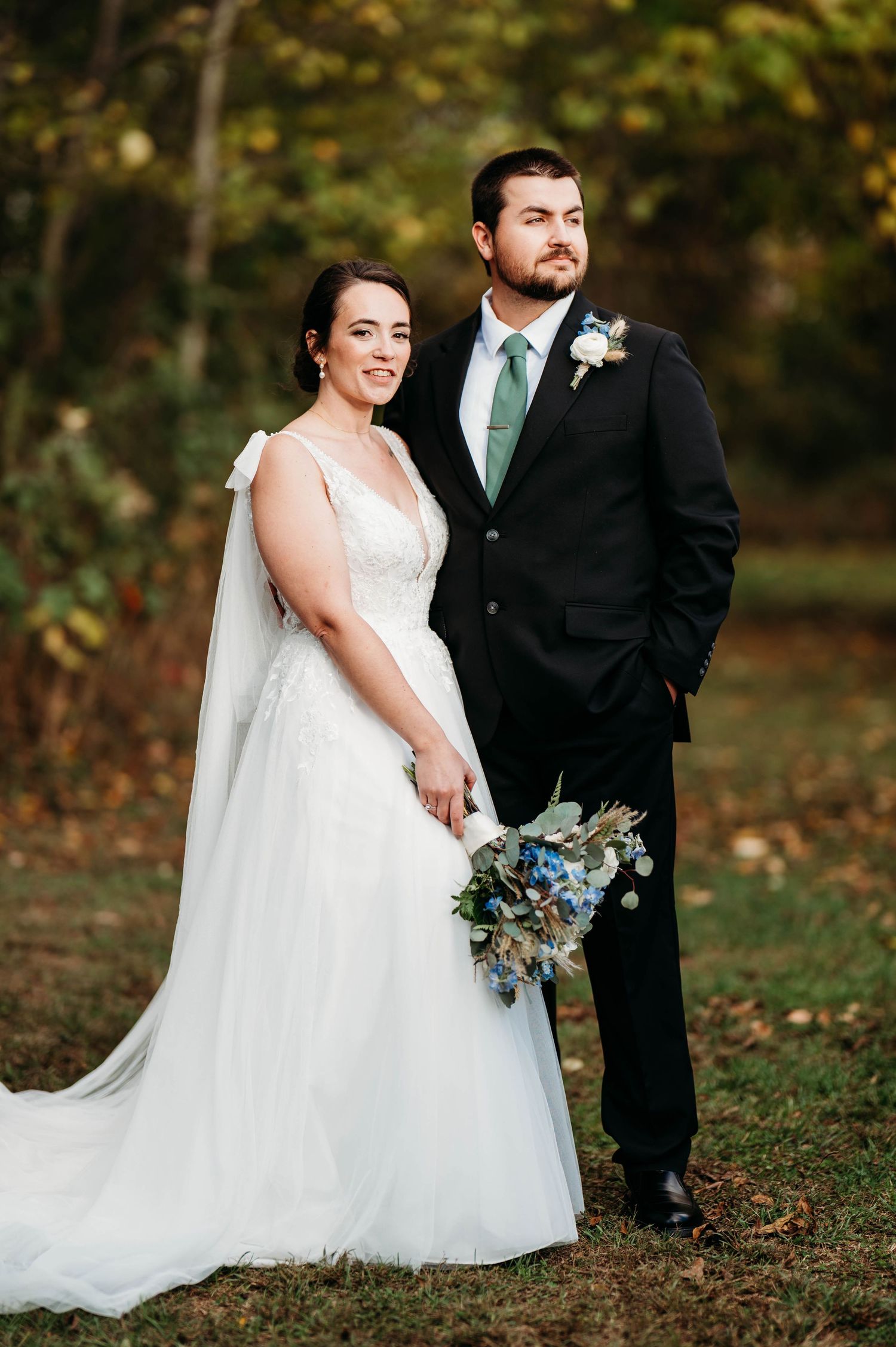 A bride and groom pose together in a wooded outdoor setting during their fall wedding ceremony.