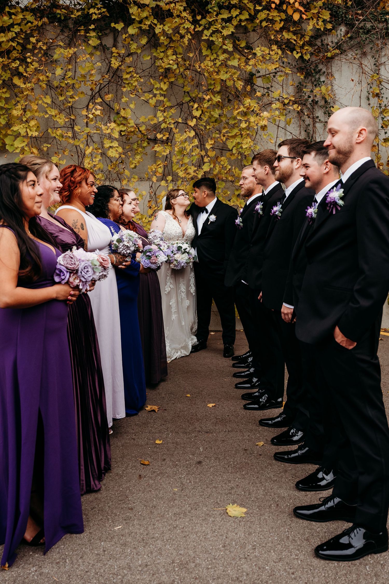 Wedding party members in purple dresses and black suits stand in two facing rows during an autumn outdoor ceremony.