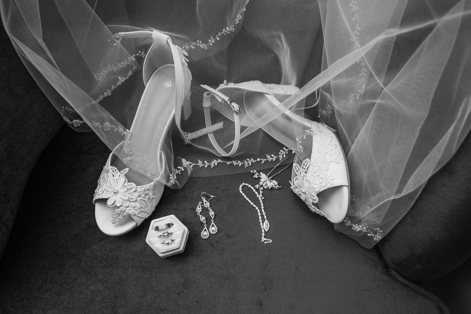 Black and white photo of bridal accessories including lace heels, earrings, necklace, and ring box beneath a flowing veil.