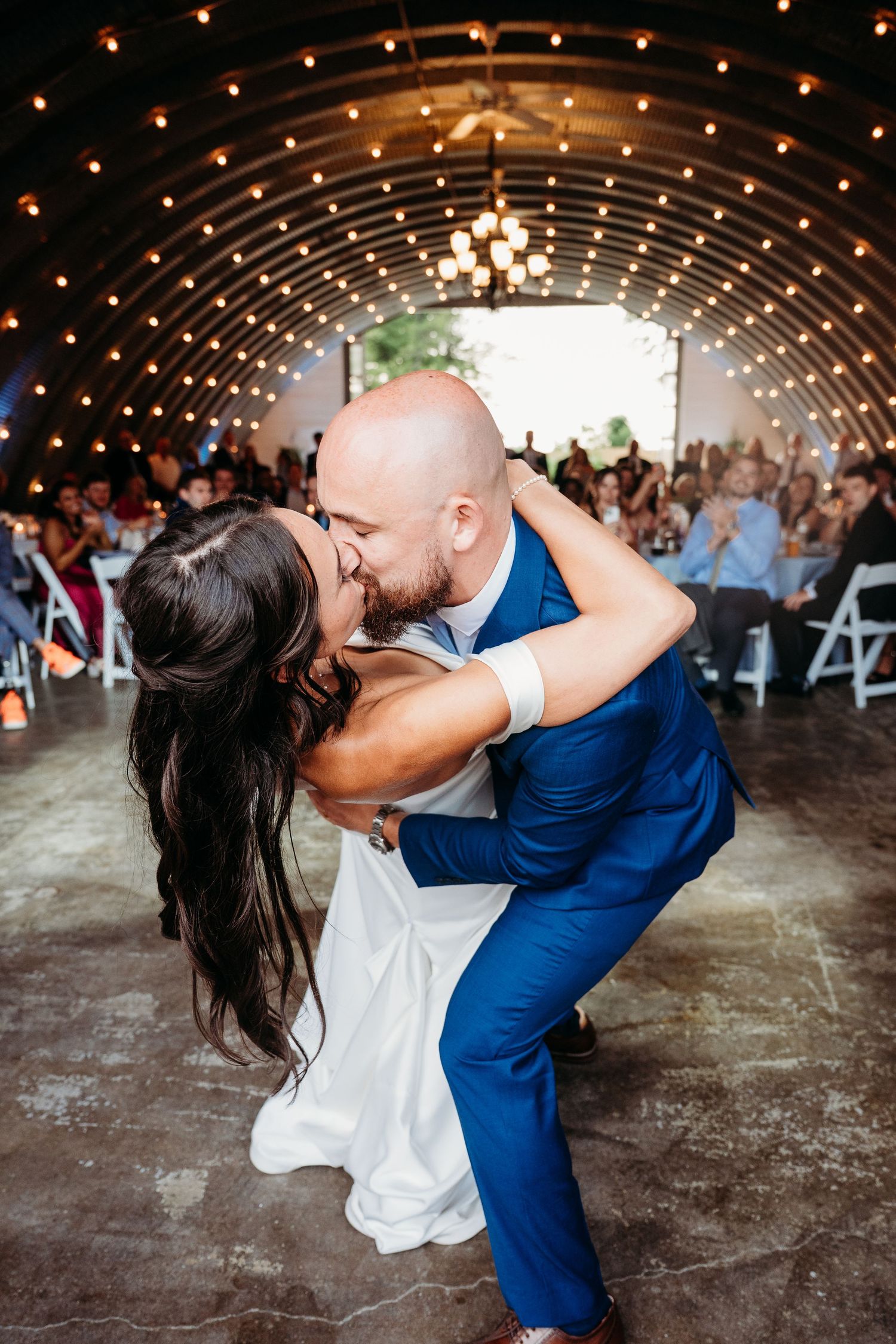 Bride and groom share a romantic dip kiss on the dance floor at their wedding reception under warm string lights.