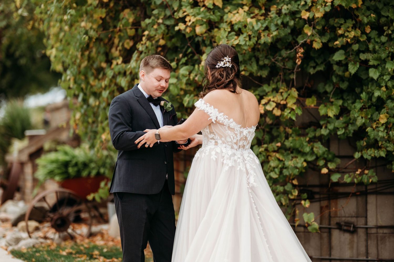 Groom in black tuxedo fastening bride's white lace wedding gown outdoors near greenery.