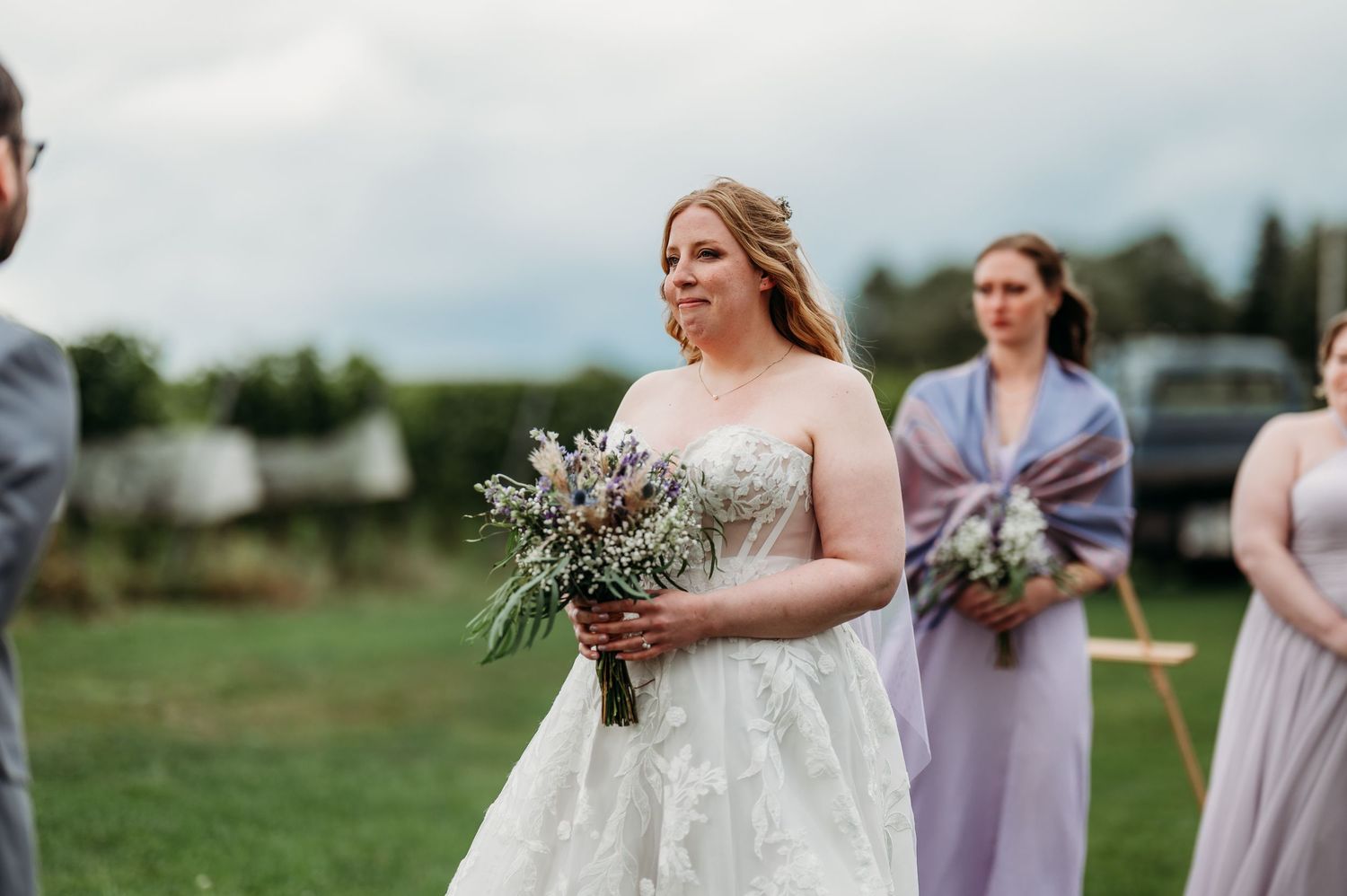 A bride and bridesmaids in lavender dresses hold bouquets while walking across a green field on a cloudy day.