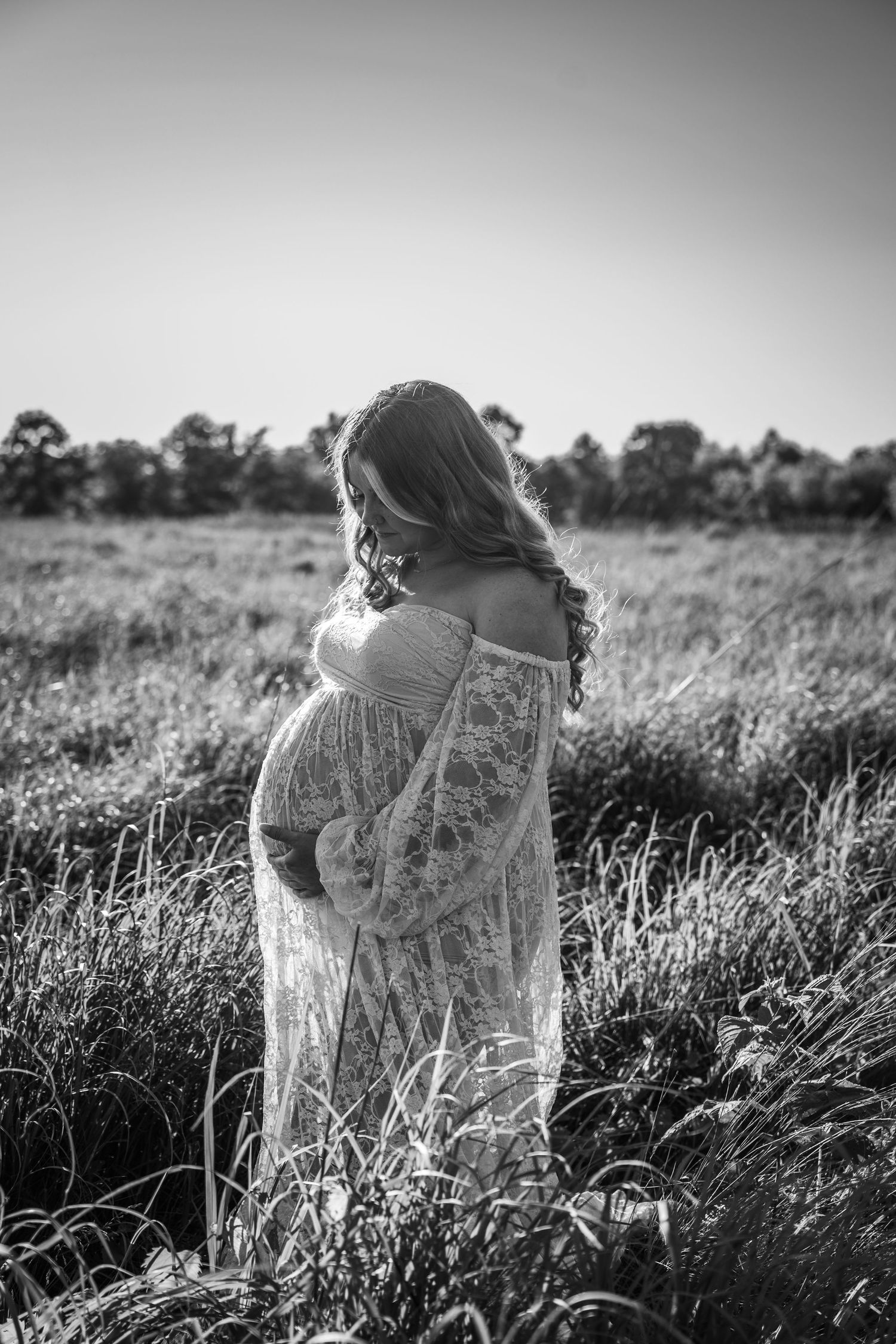 A pregnant woman in a white lace dress stands in a sunlit meadow captured in black and white photography.