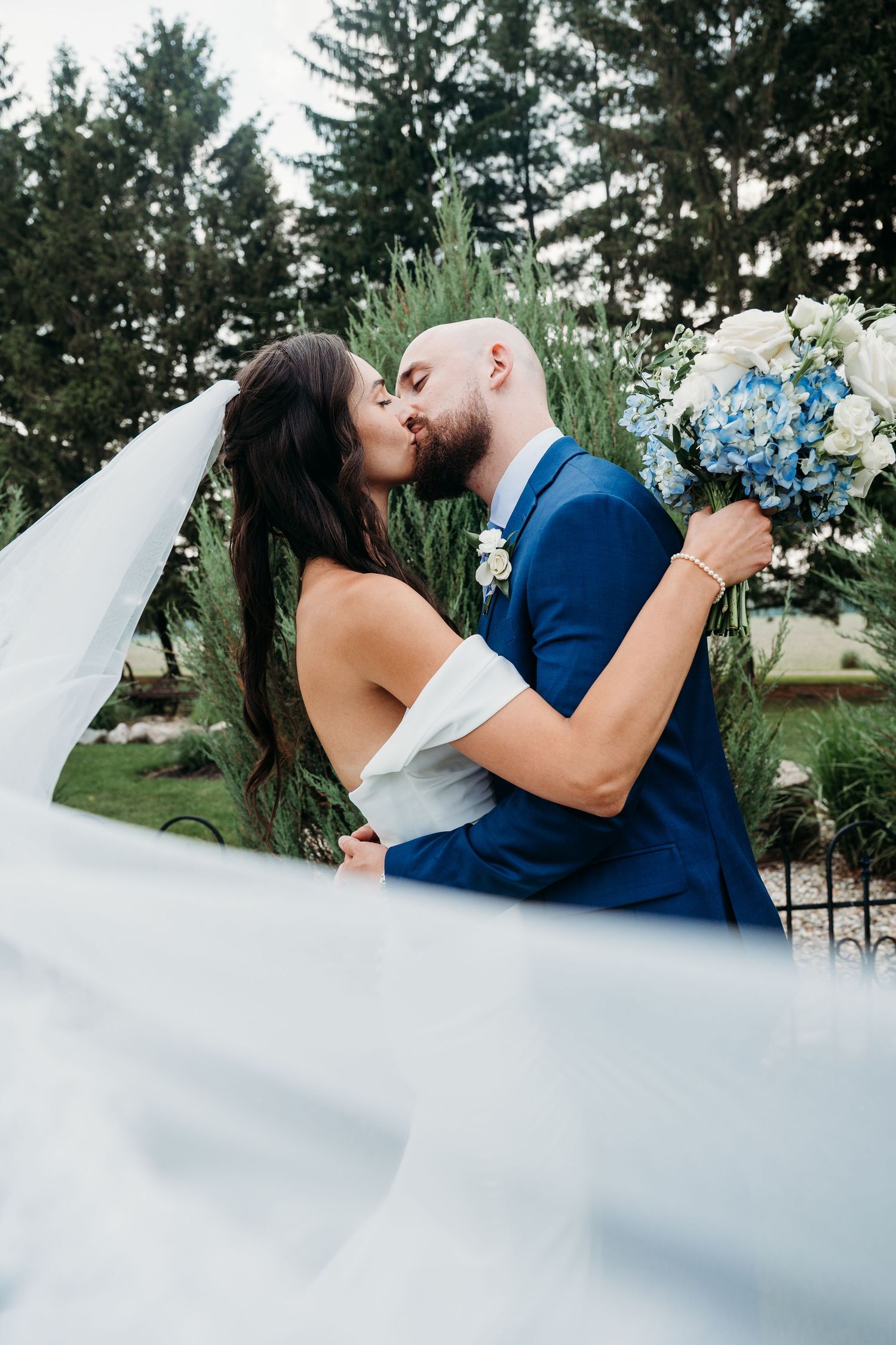 A romantic wedding couple shares a tender kiss outdoors with pine trees and a blue bouquet in their hands.