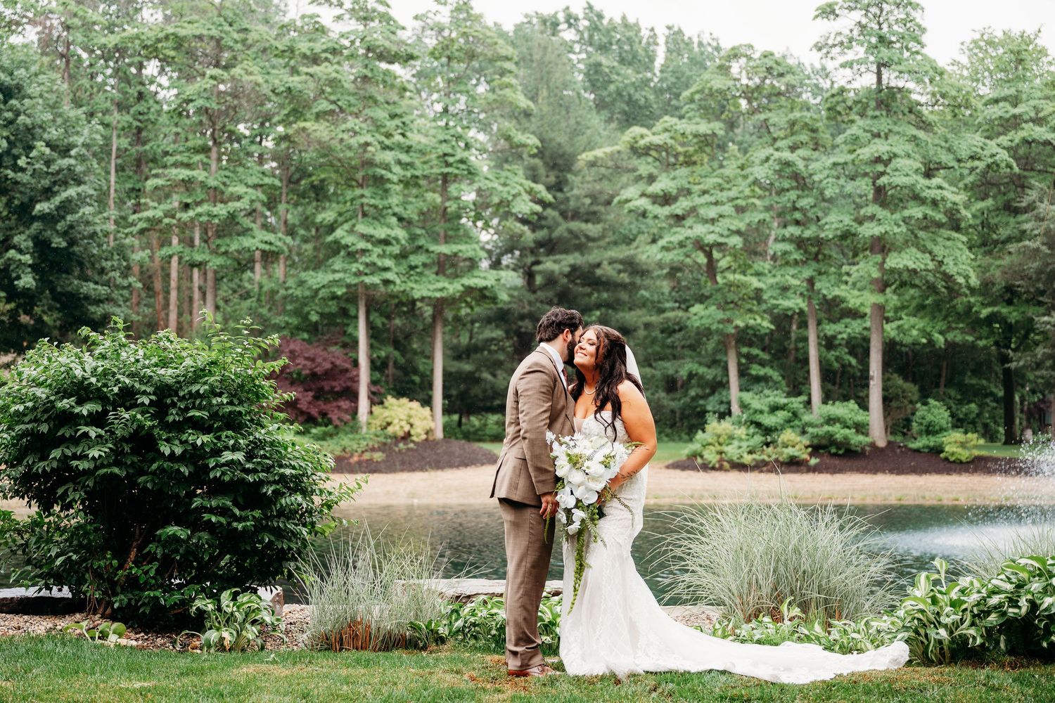 Bride and groom share a romantic kiss near a serene pond surrounded by lush green trees and gardens.