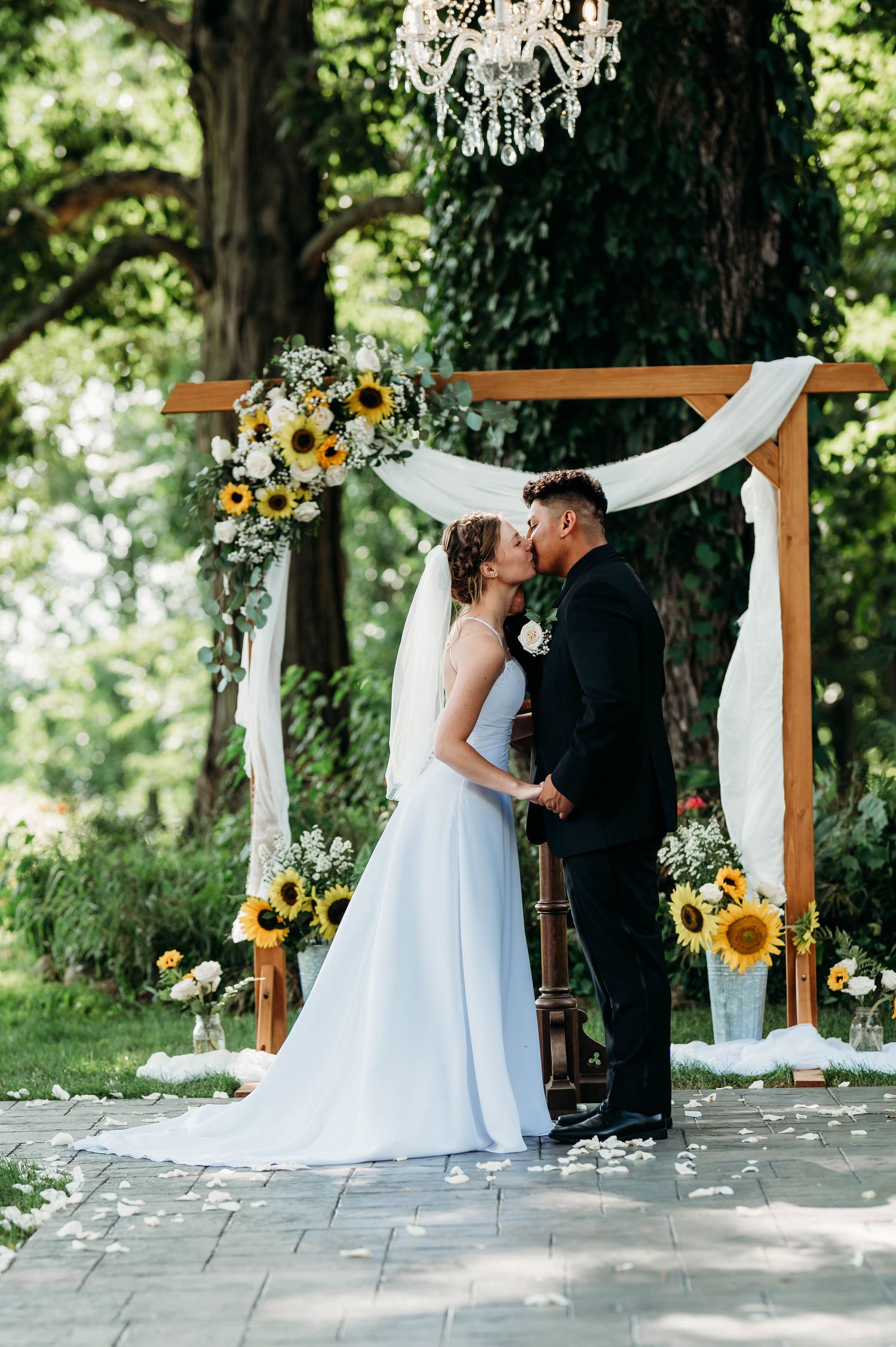 Bride and groom share first kiss under wooden arch adorned with sunflowers and white draping in outdoor garden wedding.