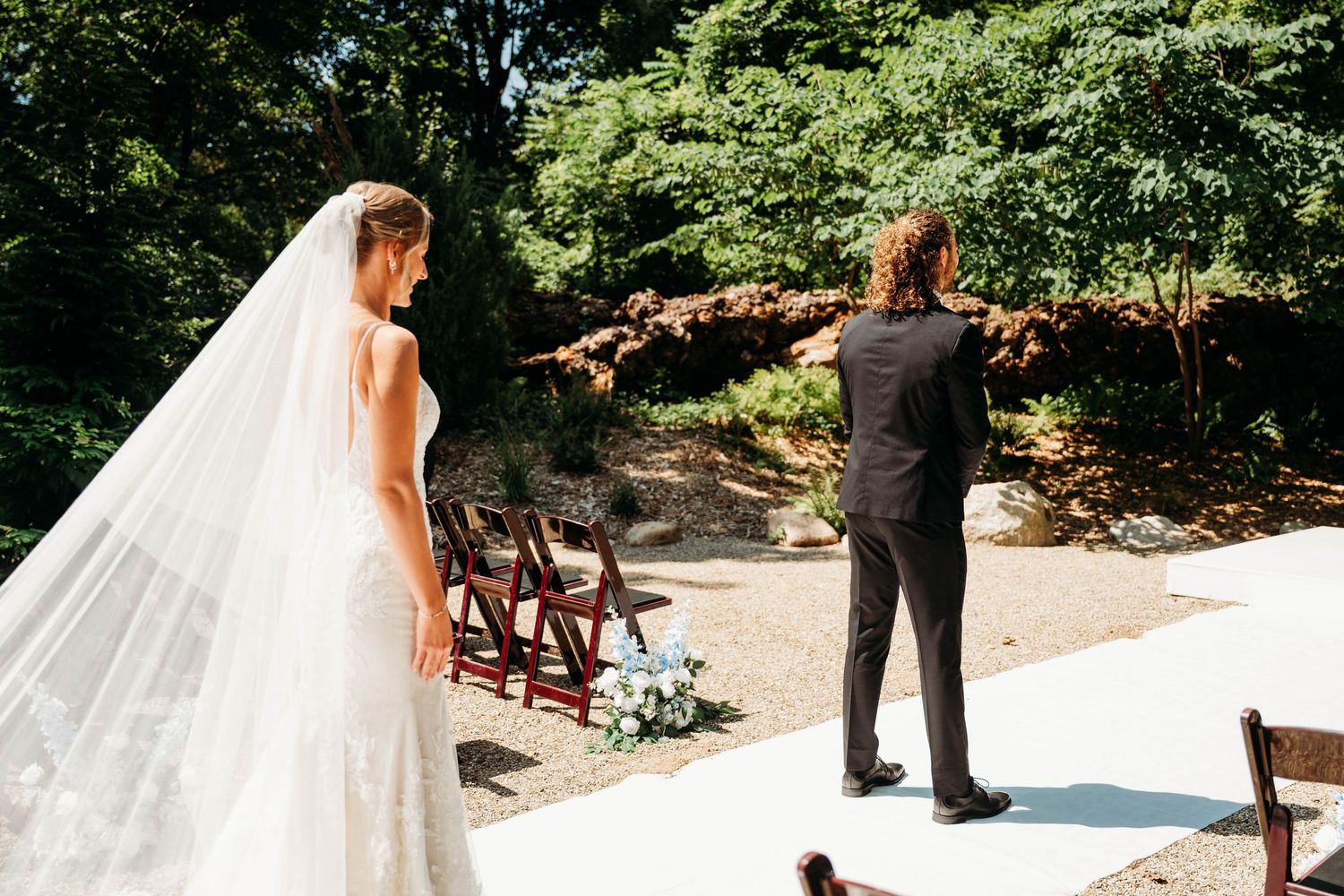 Bride in white gown and veil stands before groom in black suit at an outdoor wedding ceremony surrounded by lush greenery.
