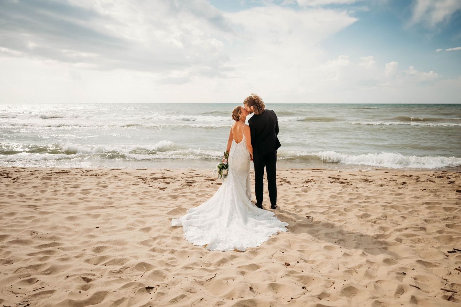 Bride and groom share a romantic kiss on a sandy beach with ocean waves in the background on their wedding day.