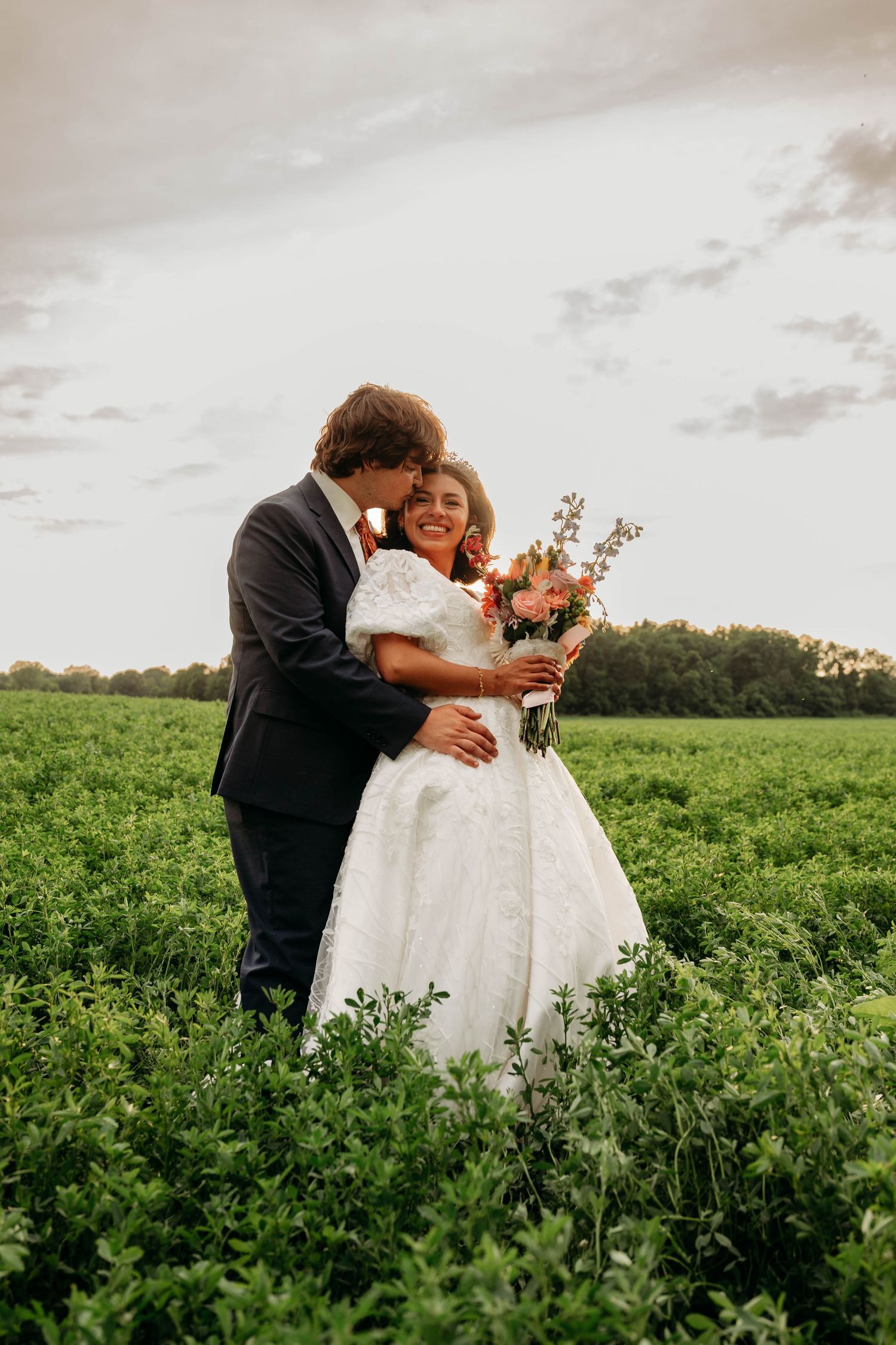 Bride and groom embracing in a lush green field, bride holding a colorful bouquet, sharing a romantic moment at sunset.
