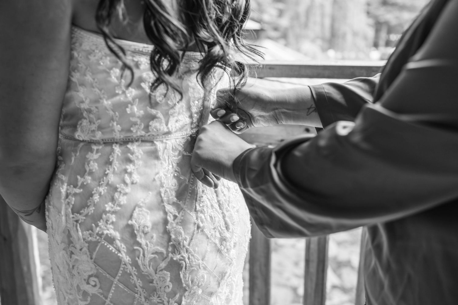 Black and white photo of hands fastening the back of a lace wedding dress.