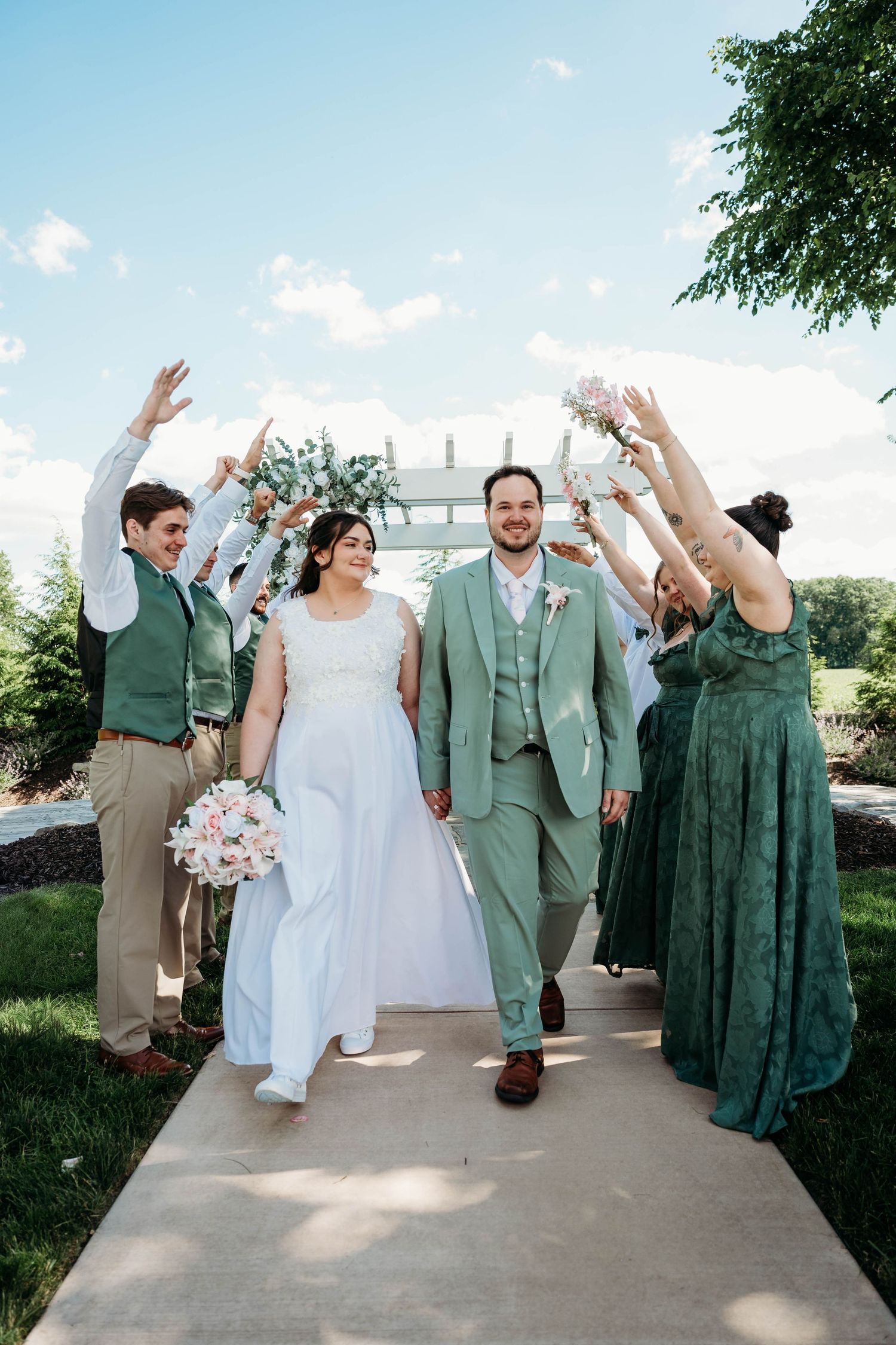 Bride and groom walk joyfully through an arch of raised hands with wedding party in sage green attire outdoors.