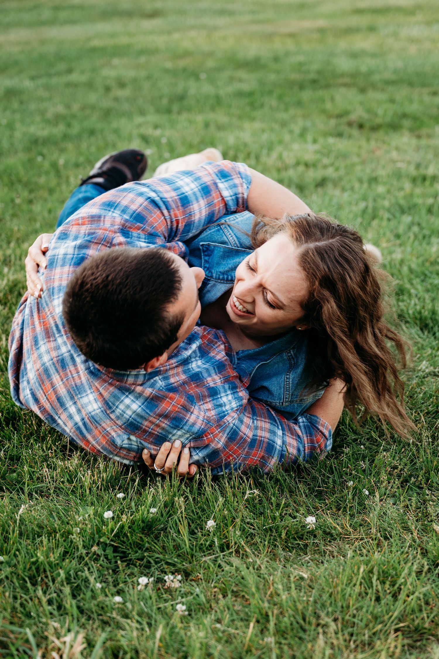 Couple laughing and cuddling together on green grass, both wearing plaid shirts on a sunny day.