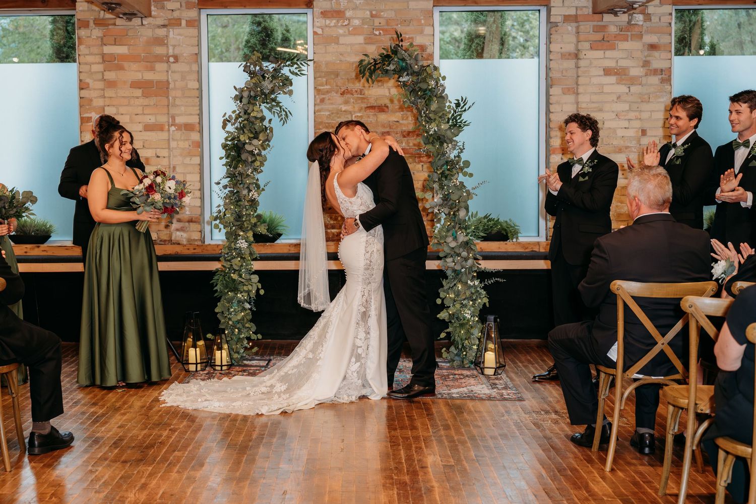 Bride and groom share first kiss at wedding ceremony with greenery arch, candles, and guests applauding in elegant venue.