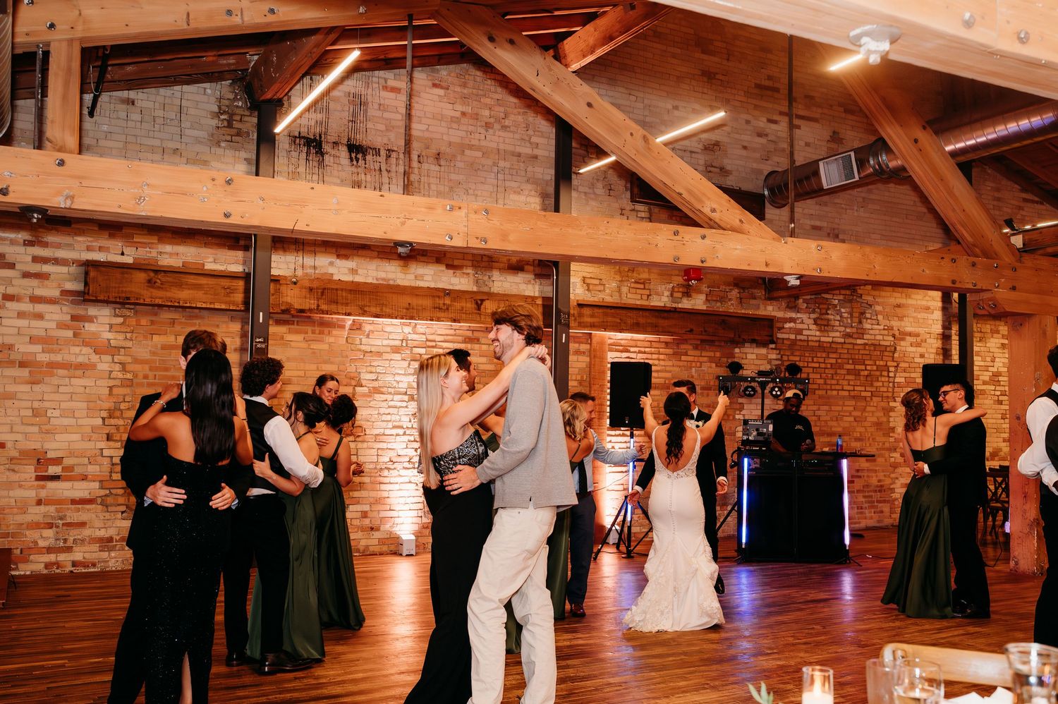 Couples dancing together at a rustic wedding reception in a barn venue with exposed brick walls and wooden beam ceiling.