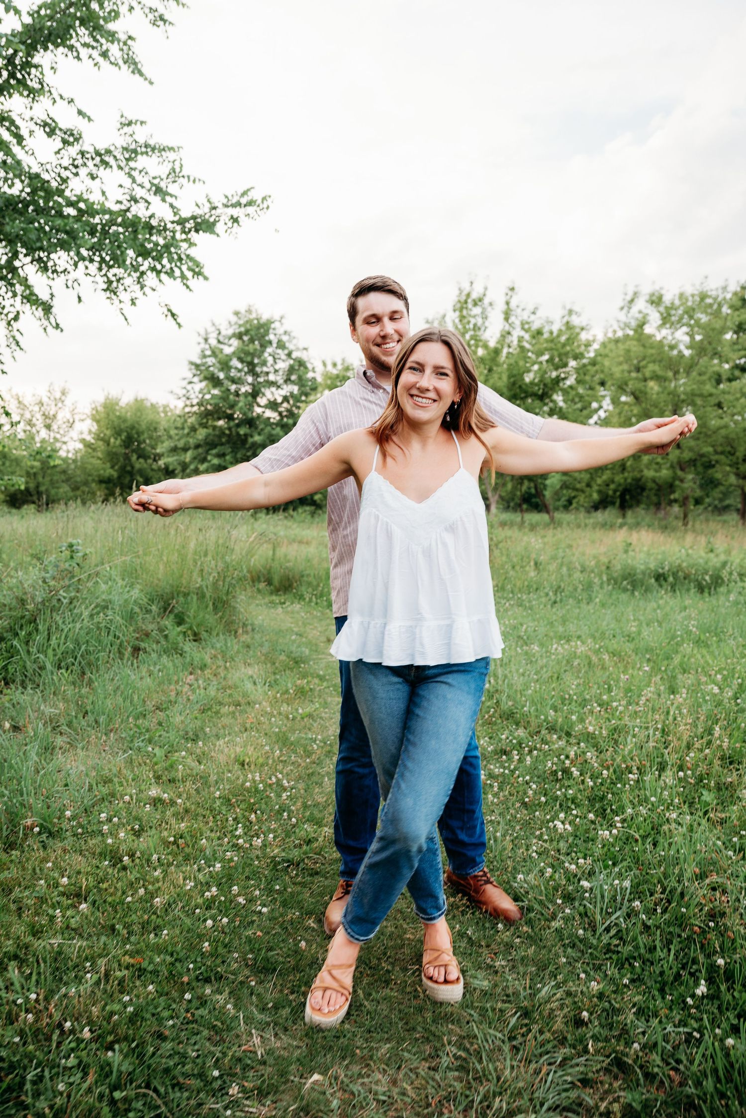 Happy couple posing outdoors in a green field, woman spreading arms wide while man stands behind her smiling.