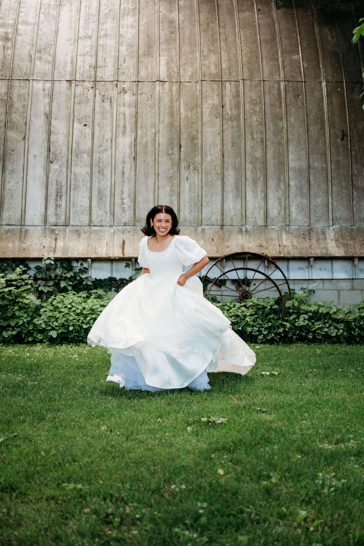 Bride in white puff-sleeve wedding gown twirling on green grass in front of rustic barn.