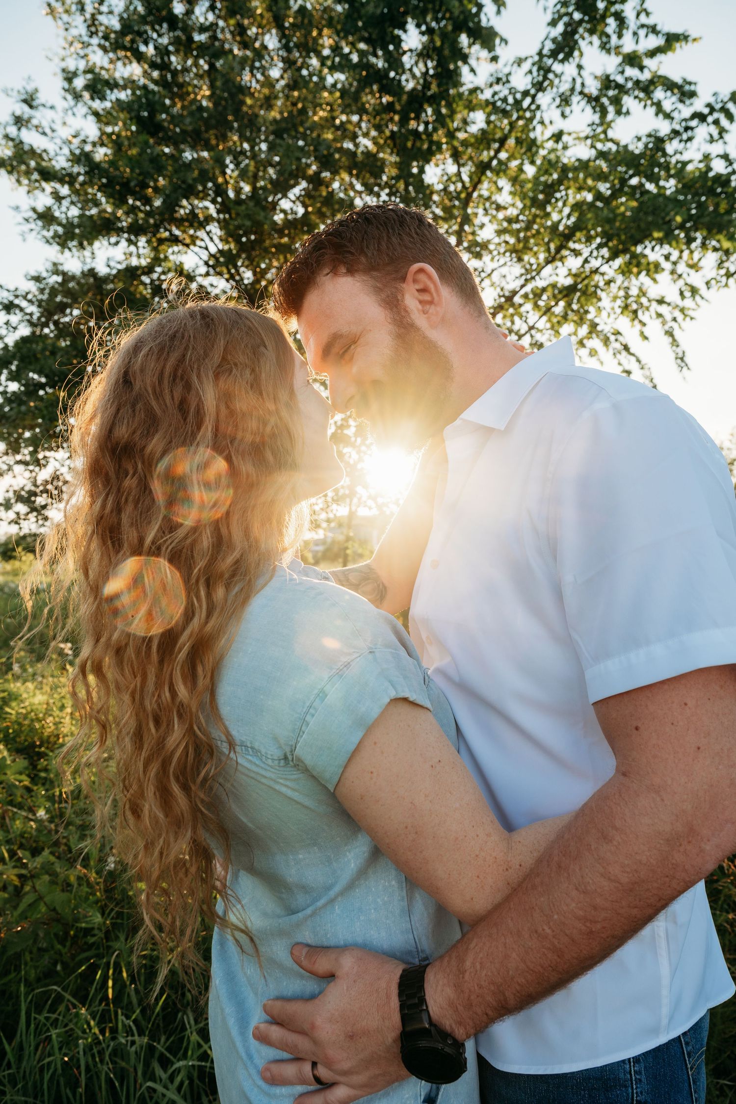 Couple embracing outdoors at golden hour with sun flare through green trees, woman in denim shirt with curly hair.