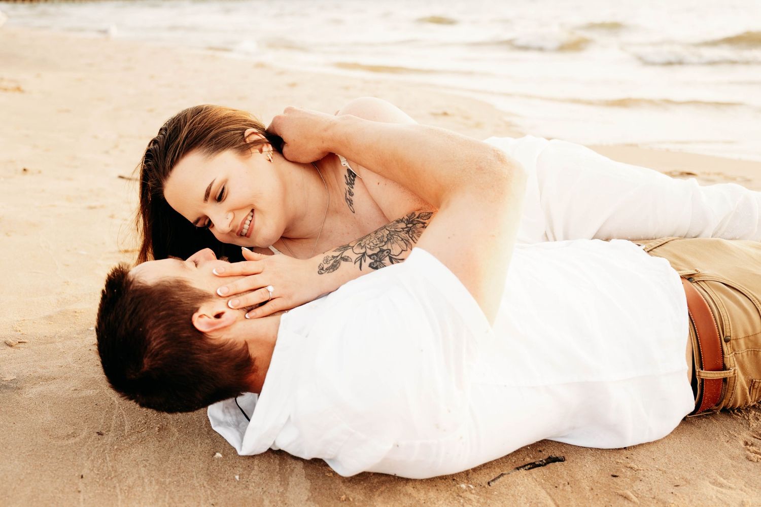 Couple sharing a romantic moment lying together on sandy beach, woman with tattoo smiling at man in white shirt.